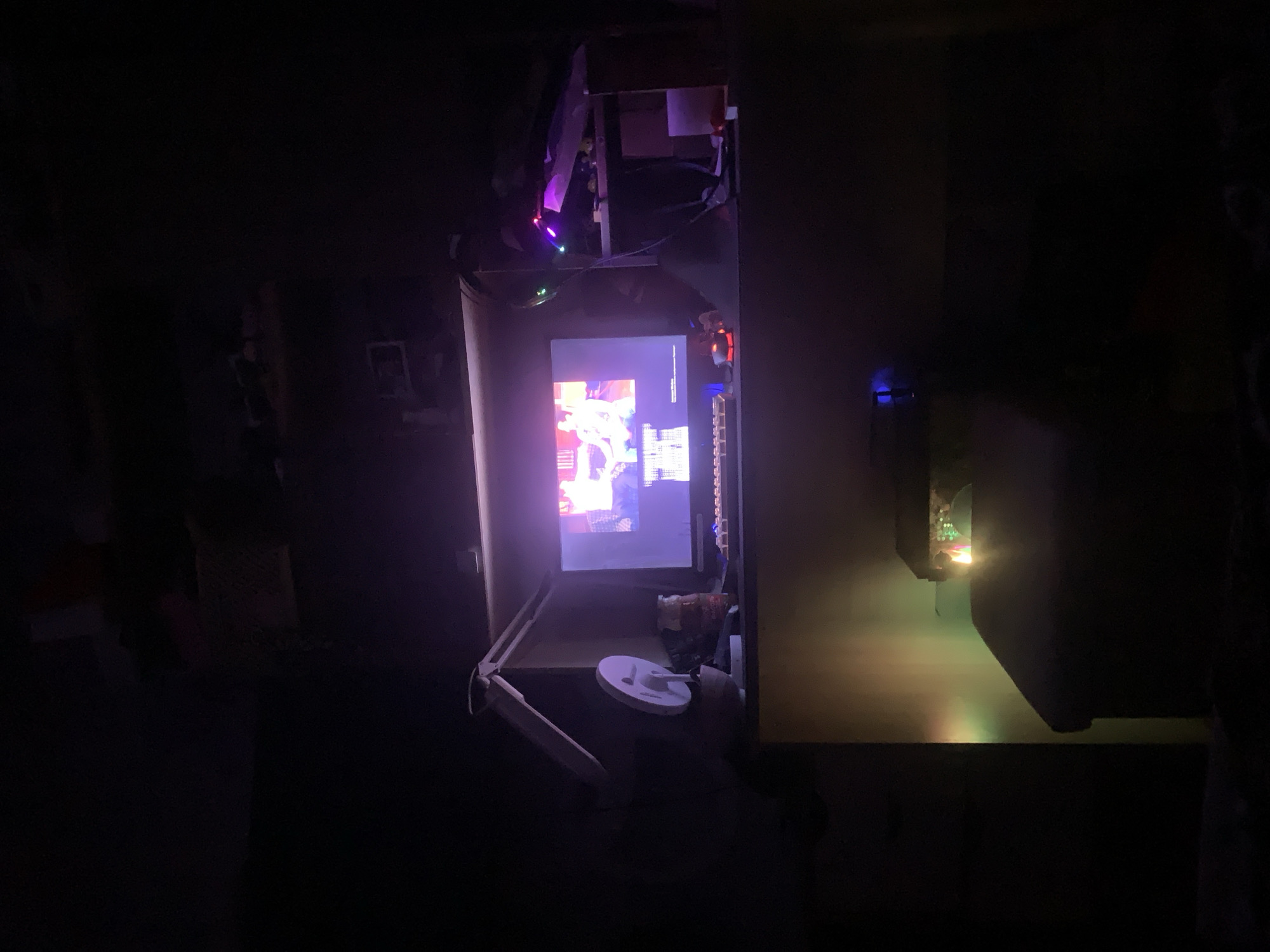In a dimly lit room, a computer monitor displays a video or image with people in it. The monitor is positioned on a desk or surface. To the left of the monitor, a white adjustable lamp is attached to the side of a cabinet. Next to the lamp, a white circular object, possibly a fan or speaker, is visible. A jar with a red lid sits on the desk in front of the monitor. To the right of the monitor, a shelf with various items, including colorful LED lights, is present. In the foreground, a dark furniture piece, likely a desk or dresser, is visible with colored lights reflecting on its surface. The overall environment suggests a personal workspace or entertainment area. The image does not contain any specific indicators that would allow for the localization of Norilsk, Russia.