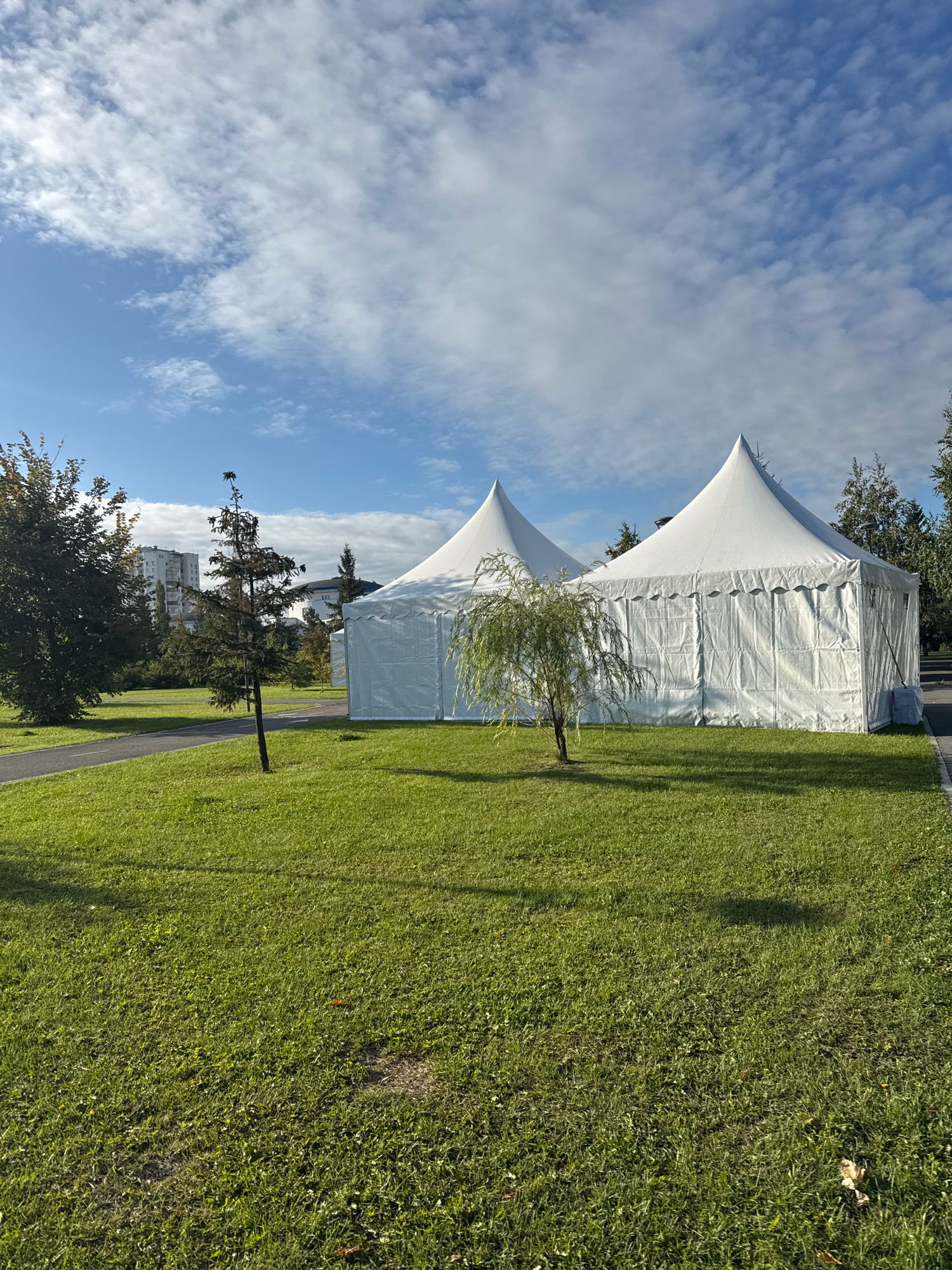 The image depicts two large white tents with pointed roofs, resembling marquees or event shelters, set up on a green lawn. The tents are located in a park-like setting. In the background, several trees are visible, along with modern apartment buildings, suggesting an urban environment. The sky is blue with scattered white clouds. The foreground is dominated by a well-maintained grassy area. There are no people visible in the image. The tents appear to be made of a white fabric material, with visible seams and tensioning elements. The overall scene suggests an outdoor event space or a temporary installation within a city park. The location is identified as being in the city of Ufa, Russia.