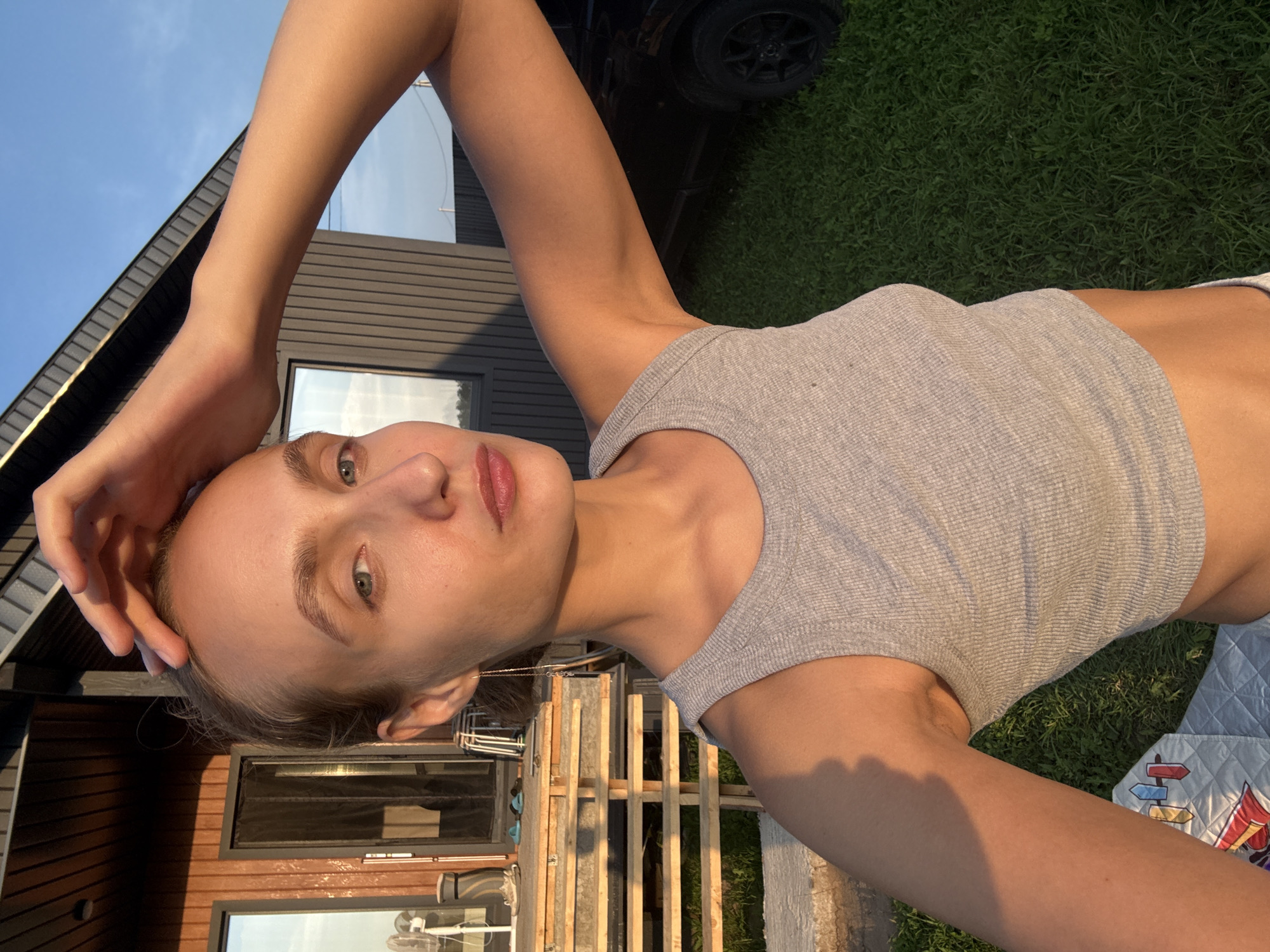 A young woman, with light skin and blonde hair tied back, is captured in a selfie taken outdoors in the village of Podshivalovo, Russia. She is wearing a light gray tank top, and her left arm is raised, with her hand resting on her head. Her face is tilted slightly towards the camera, and she has light blue eyes and pink lips. The sun appears to be setting, casting a warm golden light across the scene, suggesting it's late afternoon or early evening.
The background reveals a modern, dark-paneled building with large windows, and adjacent to it, a wooden structure made of pallets. Beyond these structures, a lush green lawn stretches out, and a portion of a dark-colored vehicle with a black wheel is visible on the grass. A quilted blanket with colorful directional signs and cartoon figures is laid out on the grass in the foreground, partially obscuring the ground. The sky above is a clear, bright blue. The overall mood seems relaxed and casual, with the golden hour lighting adding a peaceful and serene atmosphere to the setting. There is no visible text in the image.