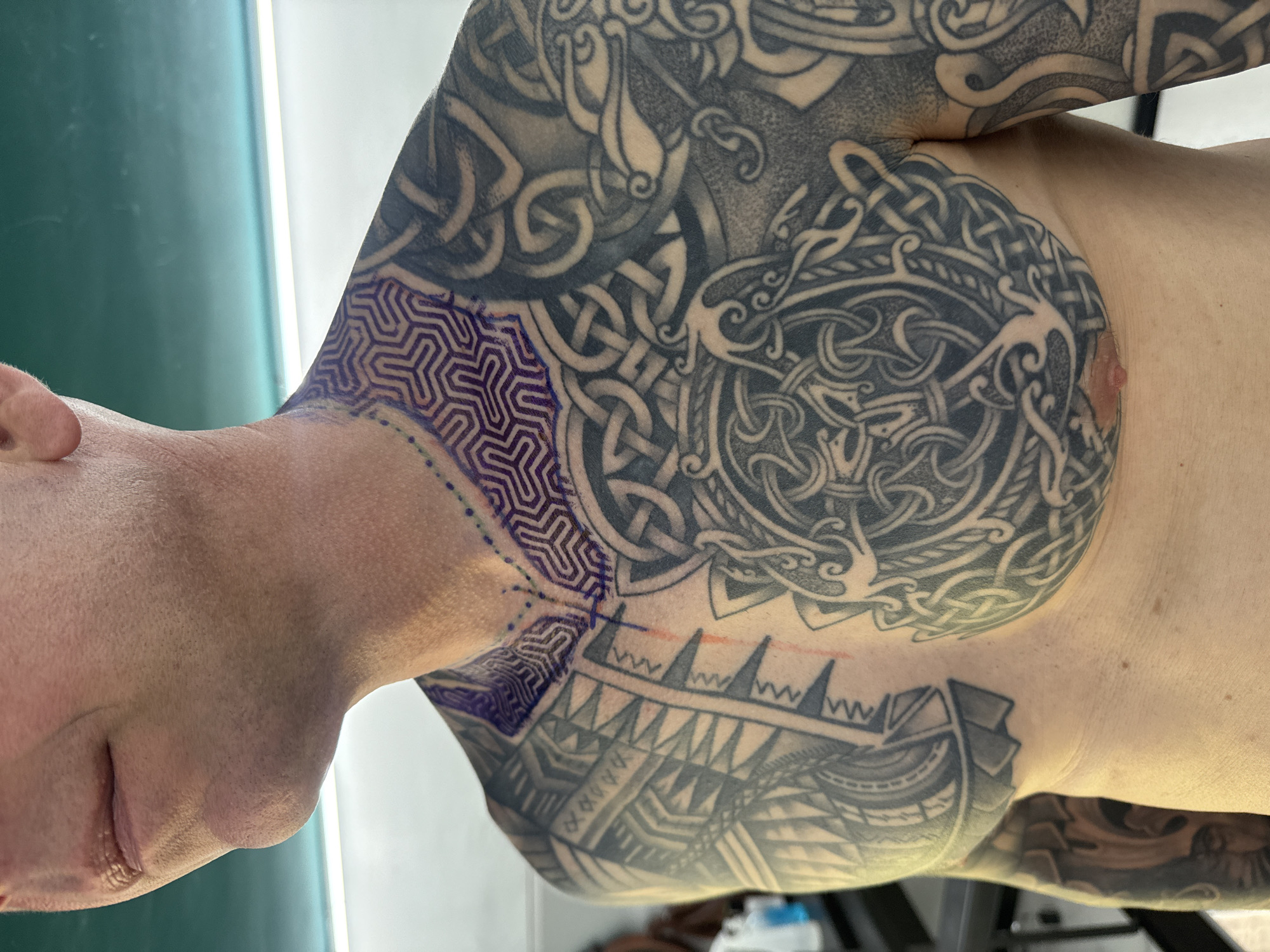 A photograph shows the upper torso and head of a light-skinned individual with extensive black ink tattoos. The tattoos cover the upper chest, shoulder, and upper arm, and they consist of intricate, interlocking patterns. The individual's eyes are closed. Blue geometric patterns are freshly drawn on the neck above the existing tattoos. There is an orange mark also on the upper chest. The background includes a dark green surface. The context suggests a tattoo studio or a setting where the individual is receiving tattoo work. The location of the scene is unknown, and the city of Sofia, Bulgaria is not discernible from the image.