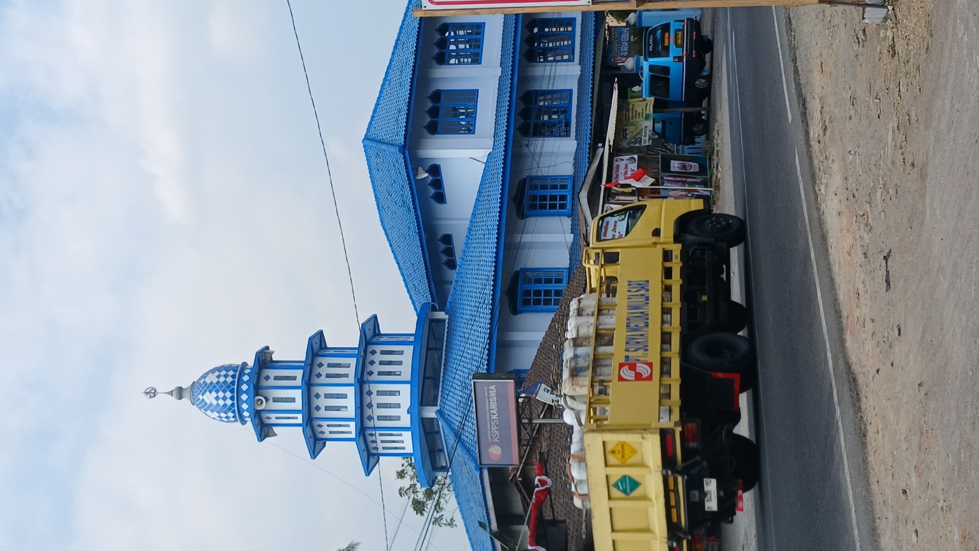 In the city of Salaman, Indonesia, a bright yellow truck with "PT SURYA MEDIKA MULIA SARI" written on its side drives past a blue and white mosque. The mosque features a prominent blue dome with white checkered patterns and several levels of windows. Adjacent to the mosque is a building with a blue tiled roof and white walls, also adorned with blue-trimmed windows. In the background, a blue minivan and other vehicles are visible, along with street signs and some vegetation. The scene is captured on a paved road under an overcast sky.