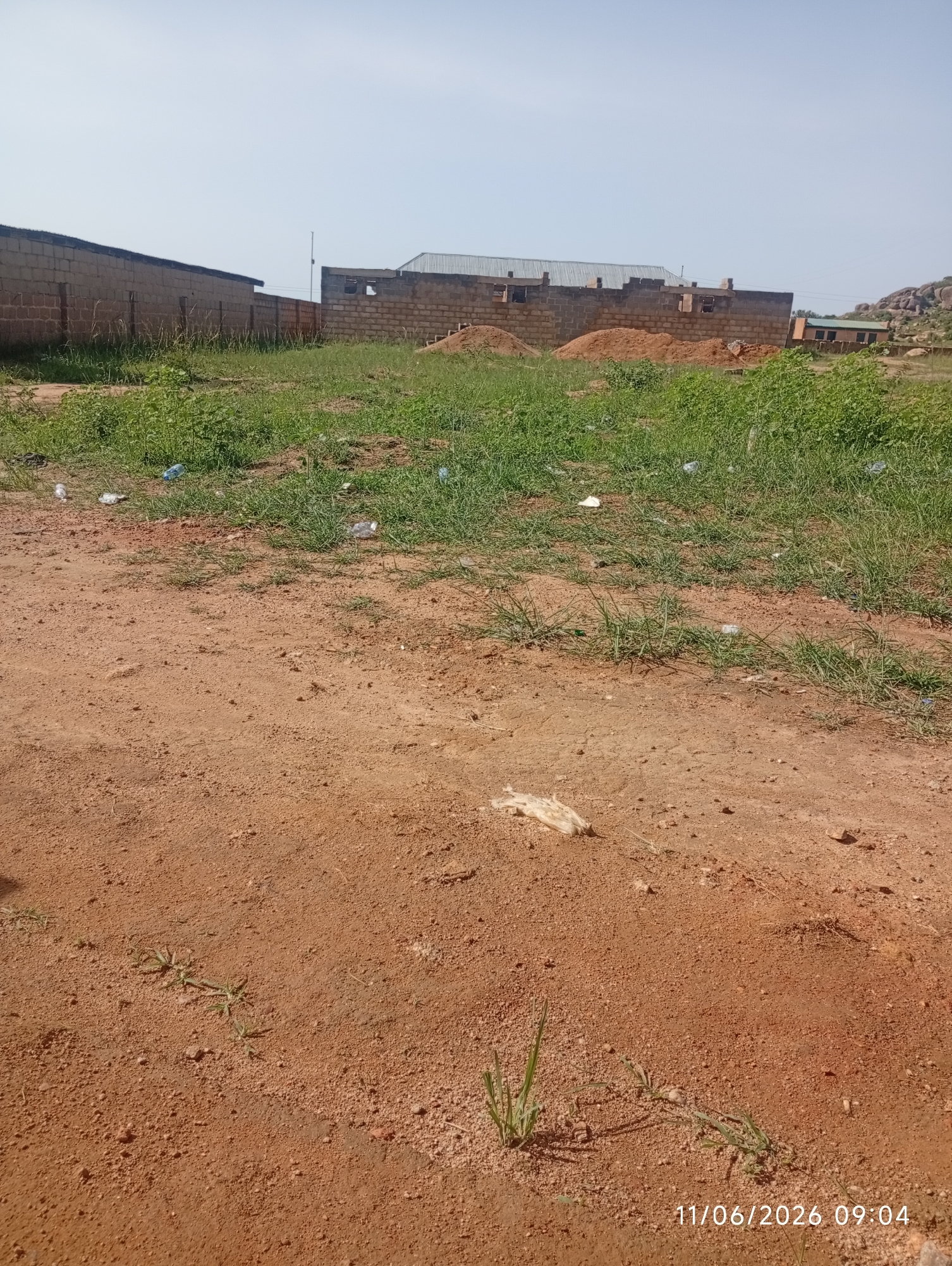 This is a medium-angle, eye-level, full, landscape photograph of a dirt and grass area in DU, Nigeria, on June 11, 2026, at 9:04 AM. In the background, there is a brick structure that appears to be under construction, with visible rebar and unfinished walls. A pile of red dirt is also present. To the left, a partially constructed brick wall stretches into the distance. The foreground is dominated by a dirt path with scattered tufts of grass and some litter, including plastic bottles. The sky is clear and bright.