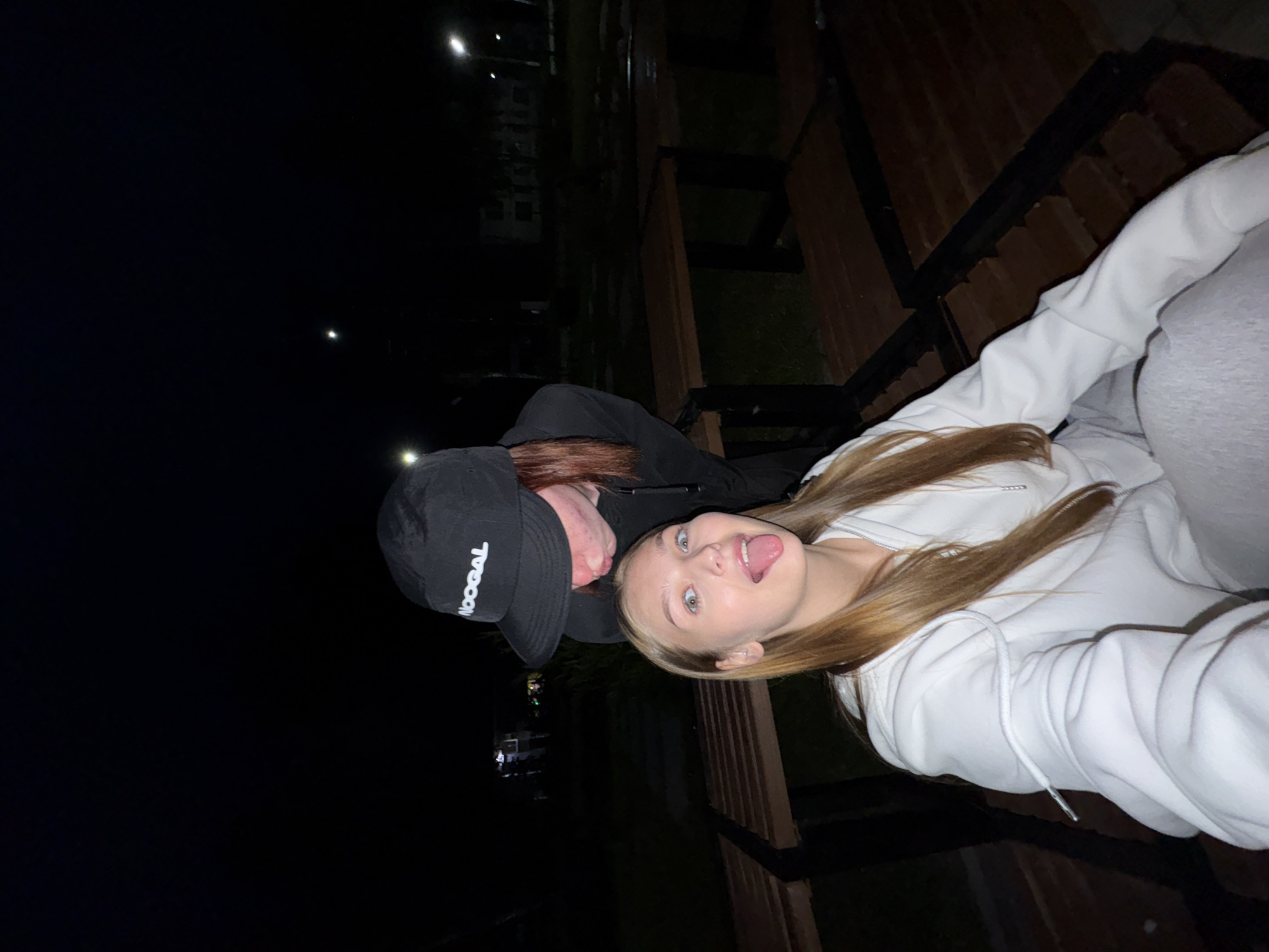 The image depicts two individuals outdoors at night, possibly in Chaikovsky, Russia. A young woman with long blonde hair is in the foreground, wearing a white hooded sweatshirt and grey sweatpants. She is sticking her tongue out, looking towards the camera with wide eyes. Behind her and to the left, a person wearing a black baseball cap with "NOOGAL" written on it and a black hoodie leans forward, with their face obscured. The background is dark, with some blurred lights and structures that could be buildings or streetlights, suggesting an urban or suburban environment. Wooden benches are visible in the mid-ground, and the ground appears to be a dark, possibly grassy or paved surface. The overall lighting is low, typical of nighttime.