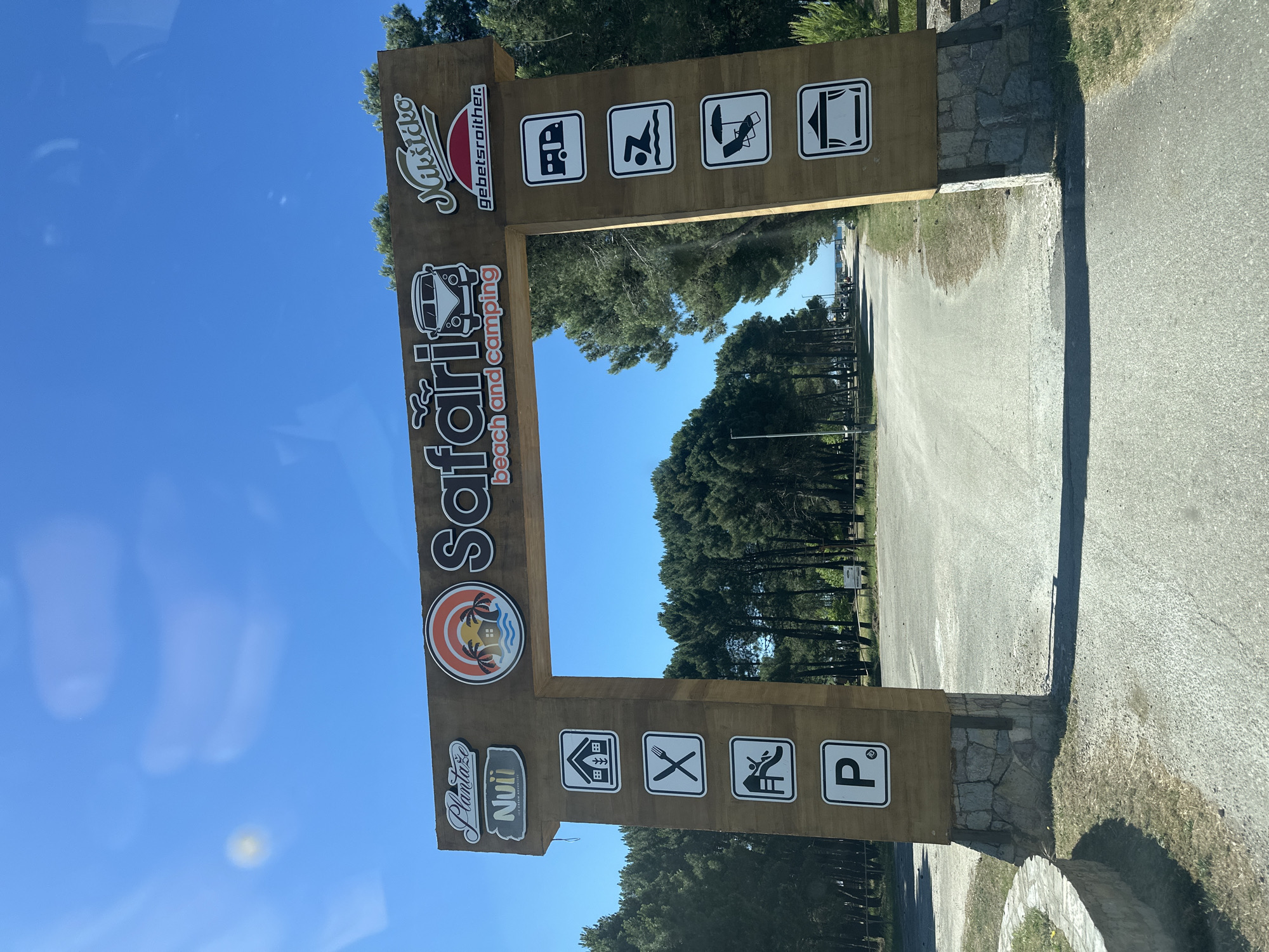 A horizontal photograph shows a sign indicating the "Safari beach and camping" location in Donji Štoj, Montenegro. The sign is made of wood and features icons and text in a stylized font. It is positioned at the entrance of a tree-lined road or path leading into the area. The day appears bright, with a clear blue sky visible between the trees. The ground is paved. The sign includes the names "Plataža Nuii" and "Ulcinjsko gebetsroither." Icons on the sign represent amenities or activities associated with the beach and camping site, such as a camper, a swimmer, a person relaxing on a beach chair, a tent, a house, food, and a parking space. The sign is supported by stone structures at its base.