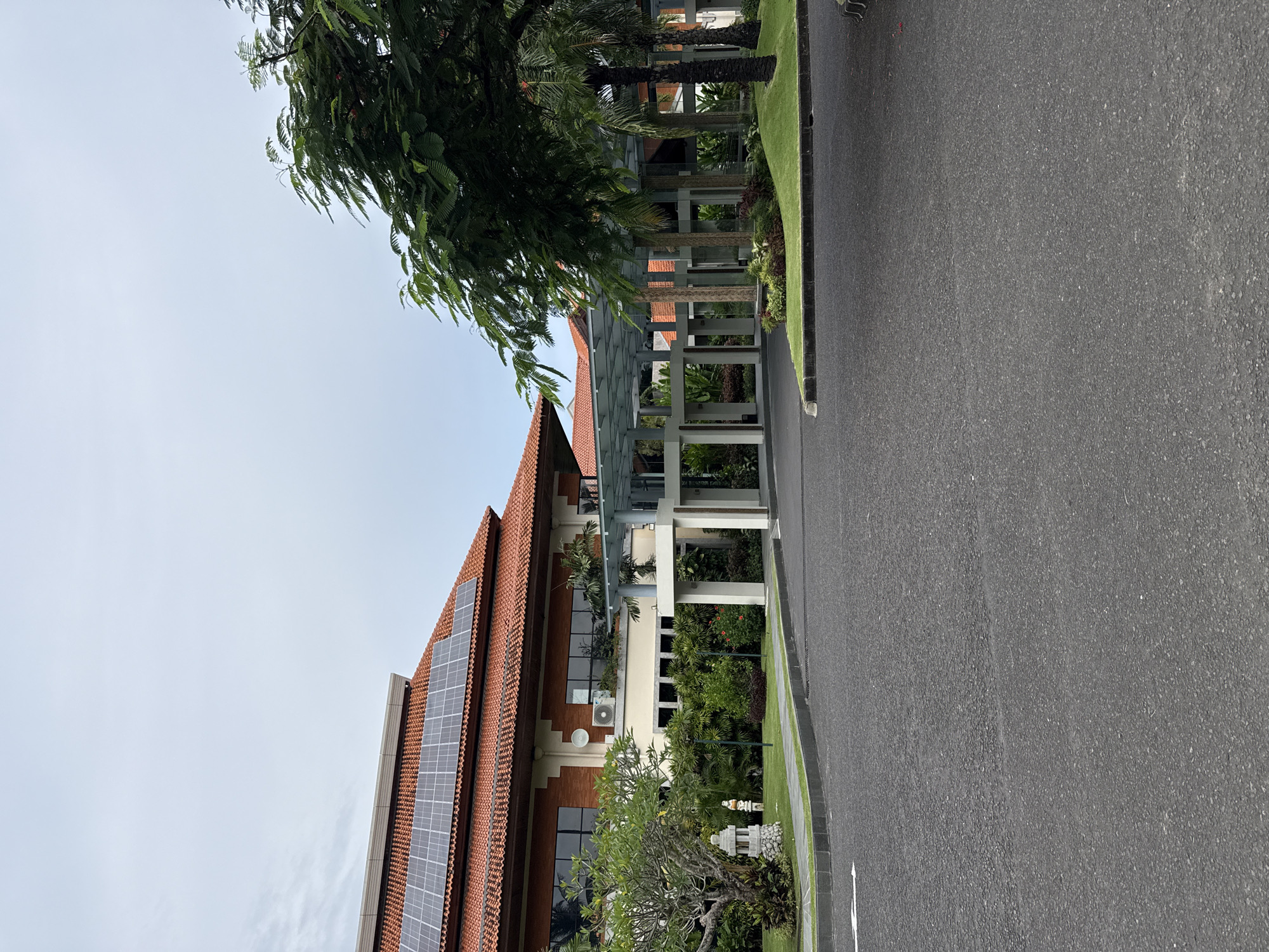 The photograph depicts a view of a building and an adjacent road, likely in Nusa Dua, Indonesia. The building features a red tile roof with solar panels installed. A covered walkway with white pillars connects to the building. Lush green vegetation is visible along the base of the building. A paved road occupies the foreground, with a narrow strip of grass and landscaping separating it from the building. The sky is overcast, suggesting a cloudy day. The scene appears to be a quiet street with a modern building integrating sustainable energy solutions.