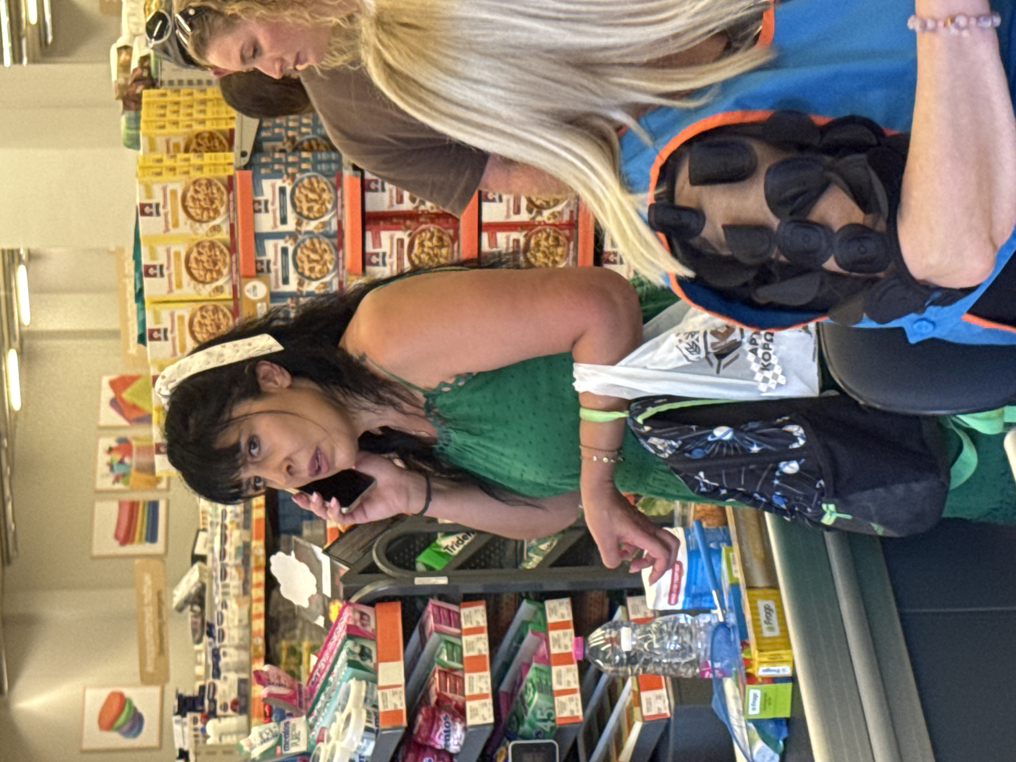 This is a medium shot, eye-level, indoor photograph taken at a checkout counter in a supermarket in Chania, Greece. The central subject is a woman of Mediterranean descent, with dark hair and bangs, wearing a green patterned sleeveless dress. She is standing and holding a smartphone to her right ear with her right hand, engaged in a phone call. Her left hand is lowered, and she is wearing several bracelets on her left wrist.
In the background, to the right of the central subject, is a woman with blonde curly hair, wearing a brown t-shirt. Her gaze is directed forward and slightly to the left. Partially visible to the right of the blonde woman is the back of another person's head, also with blonde hair.
In the foreground, on the left side of the image, is a checkout counter. On the counter, there is a clear plastic bottle of water and several items, including a yellow box with blue text that reads "Frogo". A black backpack with white and blue designs is hanging from the central subject's left shoulder. To the right, a blue vest with an orange trim is visible, worn by a person whose head is not fully visible.
The environment appears to be a retail store with shelves stocked with various products, including boxes of cereal visible in the background. The lighting is bright and artificial, typical of an indoor commercial setting.