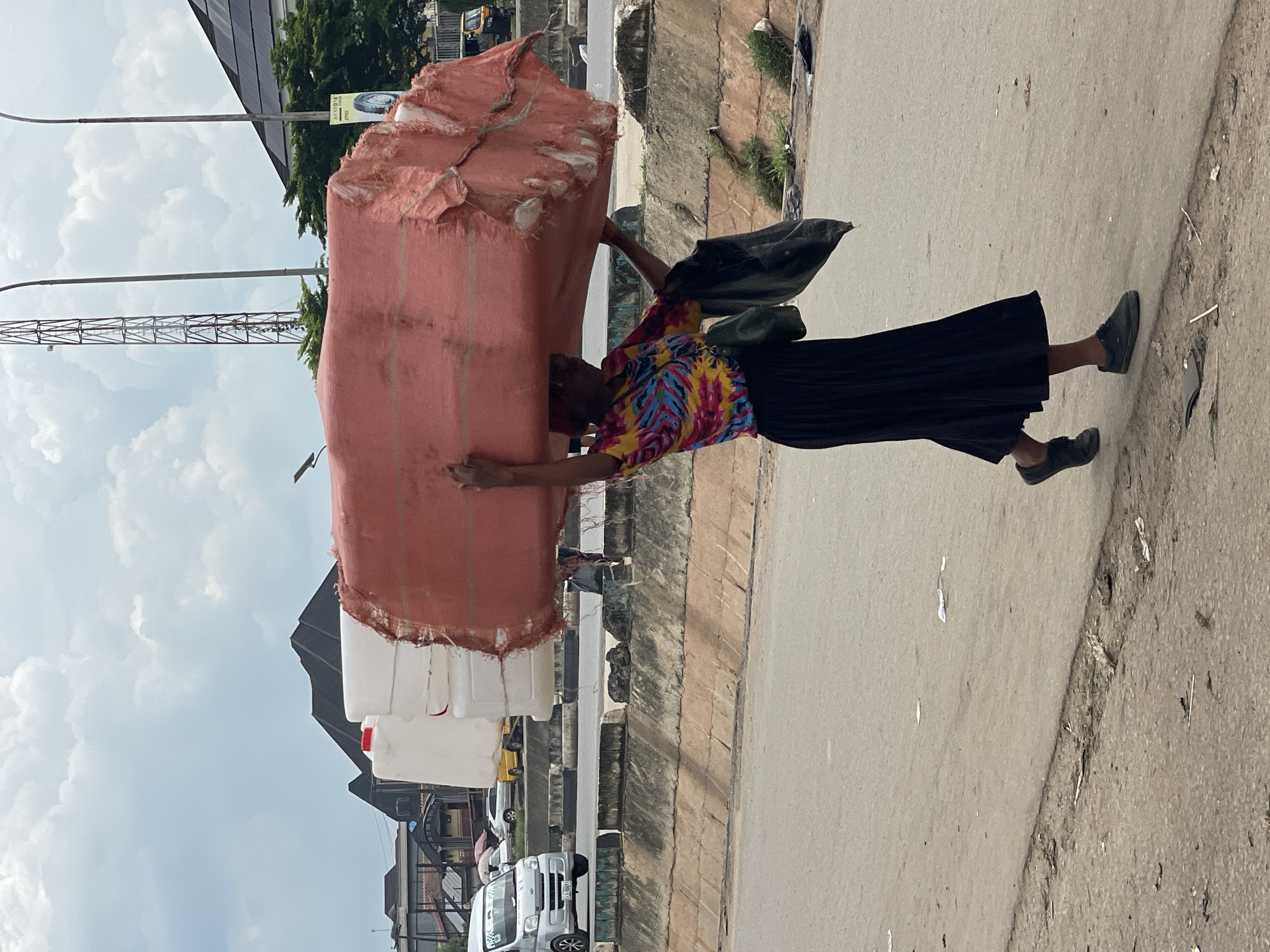 A woman is walking on a street, carrying a large bundle of white plastic containers wrapped in a reddish-brown fabric on her head. She is wearing a colorful, tie-dye t-shirt, a long black skirt, and dark shoes. A green bag hangs from her shoulder, and she holds a black plastic bag in her left hand. The street appears to be in an urban area with a concrete barrier to the left, separating the road from a dirt embankment with some sparse vegetation. In the background, there are buildings, a traffic light, a communication tower, and several vehicles, including a white van, a yellow car, and a three-wheeled vehicle. The sky is cloudy.