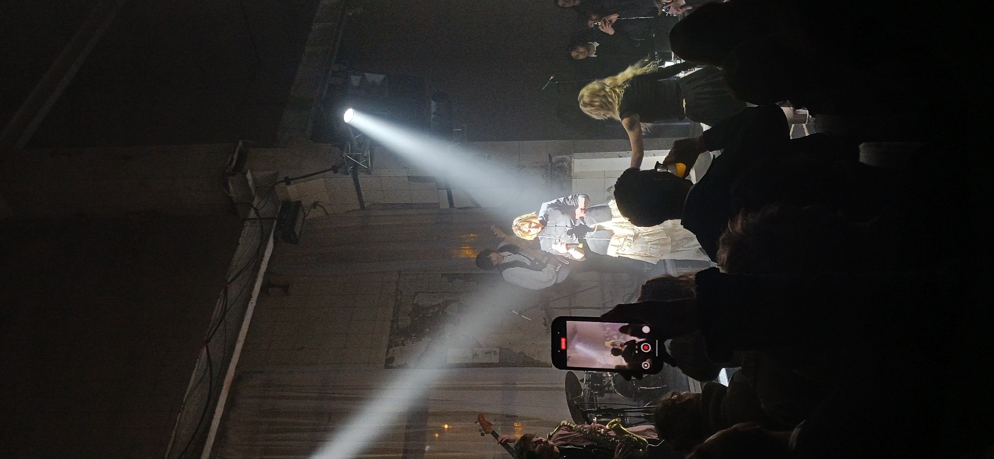 A low-angle, indoor shot, possibly a concert or performance space, taken in Unknown, Unknown. The stage is illuminated by a spotlight, capturing musicians performing. In the foreground, spectators are observing the event, with one person visible holding a smartphone to record the performance. The architectural details of the venue are partially visible, suggesting an aged or unconventional structure.