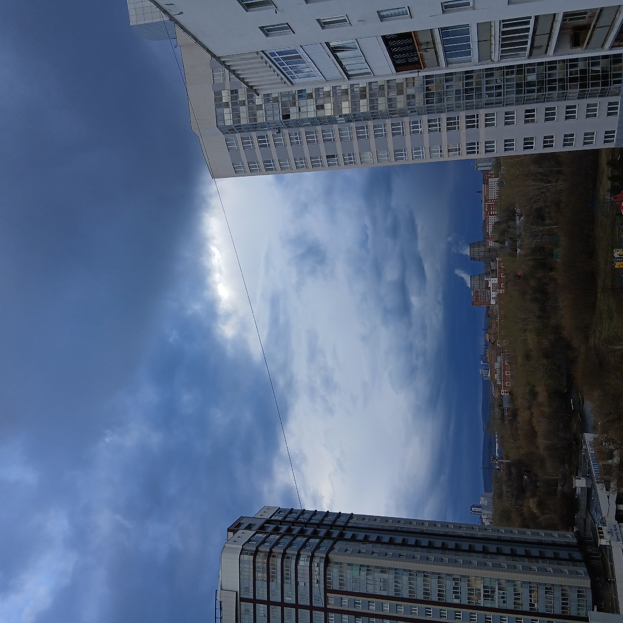 This image captures a dynamic cityscape under a dramatic sky, likely in Yekaterinburg, Russia. The perspective is from an elevated position, looking out between two modern, multi-story residential buildings that frame the view on the left and right. These buildings are predominantly light grey or white, featuring numerous windows and glass balconies.
The sky is the most striking element, showcasing a stark contrast in weather. On the left side, dark, deep blue, heavy clouds dominate, suggesting an impending storm or dense overcast conditions. Towards the center and right, the sky transitions dramatically to a lighter blue with bright, wispy white clouds, indicating a break in the weather or a different cloud system. A thin, dark utility line stretches horizontally across this changing sky.
Below the sky, a band of bare deciduous trees, possibly on a hillside or embankment, forms a natural division. Beyond the trees, a distant urban landscape is visible, with various buildings of different architectural styles, some appearing older or industrial, extending towards a hazy horizon.
There is no discernible human activity. The scene is static, capturing a moment in time. The weather appears to be in transition, likely during daytime. On a white sign at the base of a building in the distant cityscape, Cyrillic text is faintly visible, reading "Центр Коли" (Tsentr Koli).