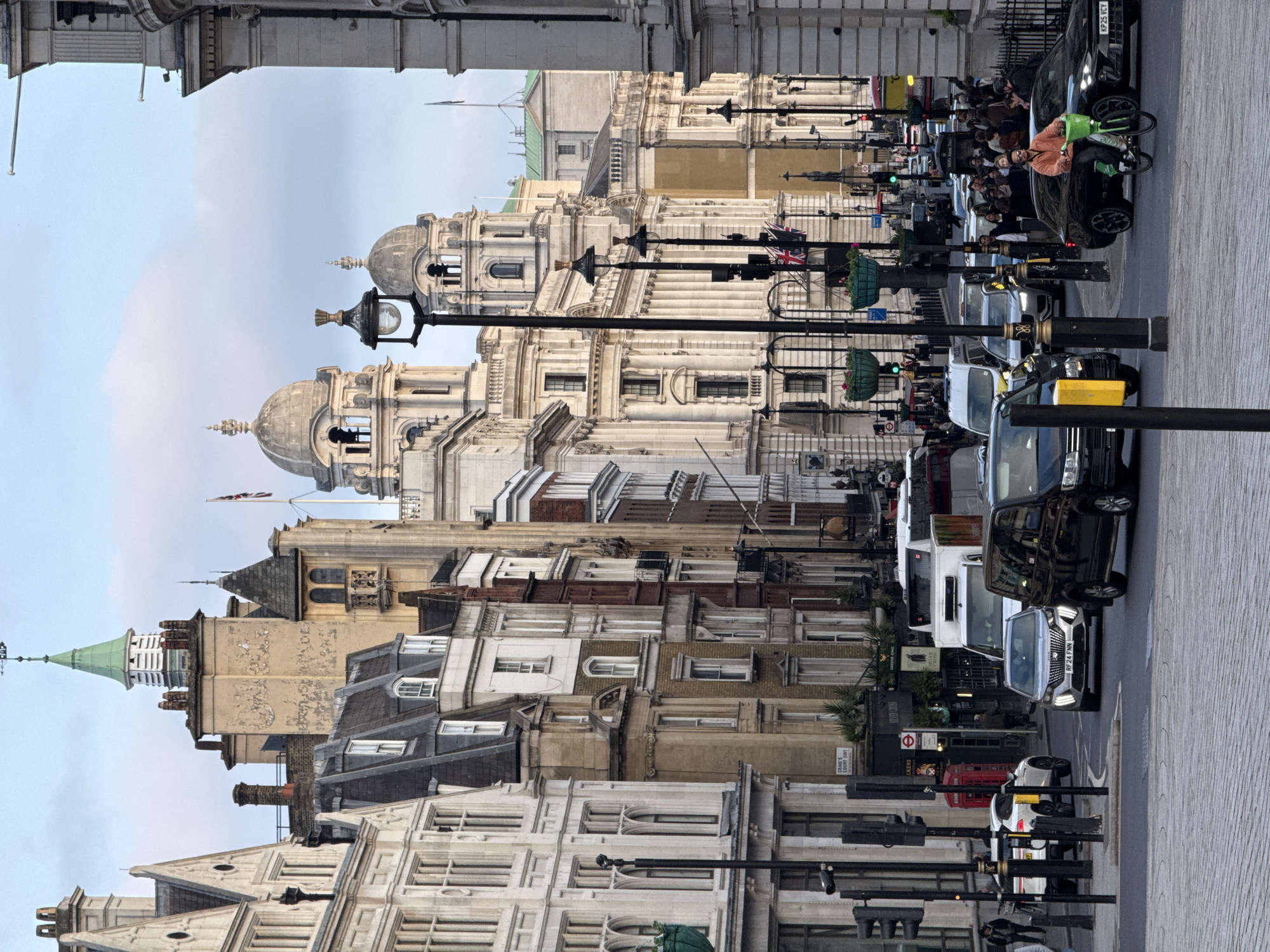 This is a street scene in London, United Kingdom. The camera is positioned at street level, capturing the buildings and traffic on a street. The architecture features a mix of historic buildings with white stone facades, decorative domes, and various architectural styles. In the lower portion of the view, there are multi-story structures with windows and varying rooflines. A green-roofed spire is visible among the buildings.
The street is populated with cars, vans, and pedestrians. A crowd of people are near parked vehicles on the right side of the image. There are also traffic lights and street lamps along the roadside. The sky is overcast.