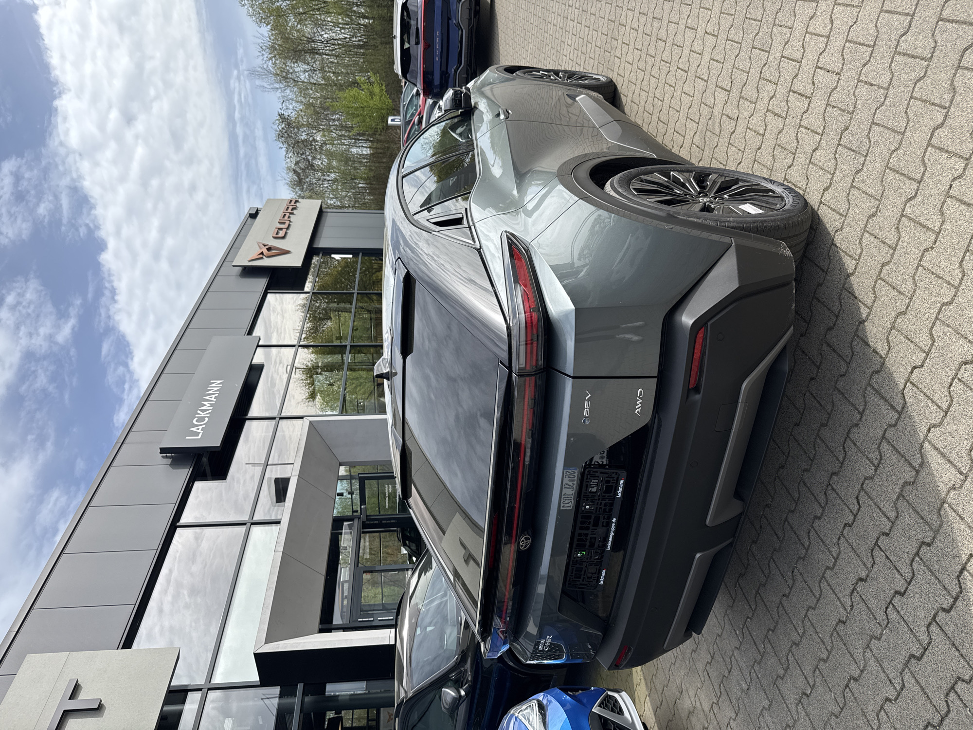 The image shows a gray Cupra Tavascan BEV AWD parked in front of a Lackmann dealership. The dealership appears to be located in Solingen, Germany, based on the license plate frame. Other vehicles are parked nearby. The image is taken outdoors on a sunny day.