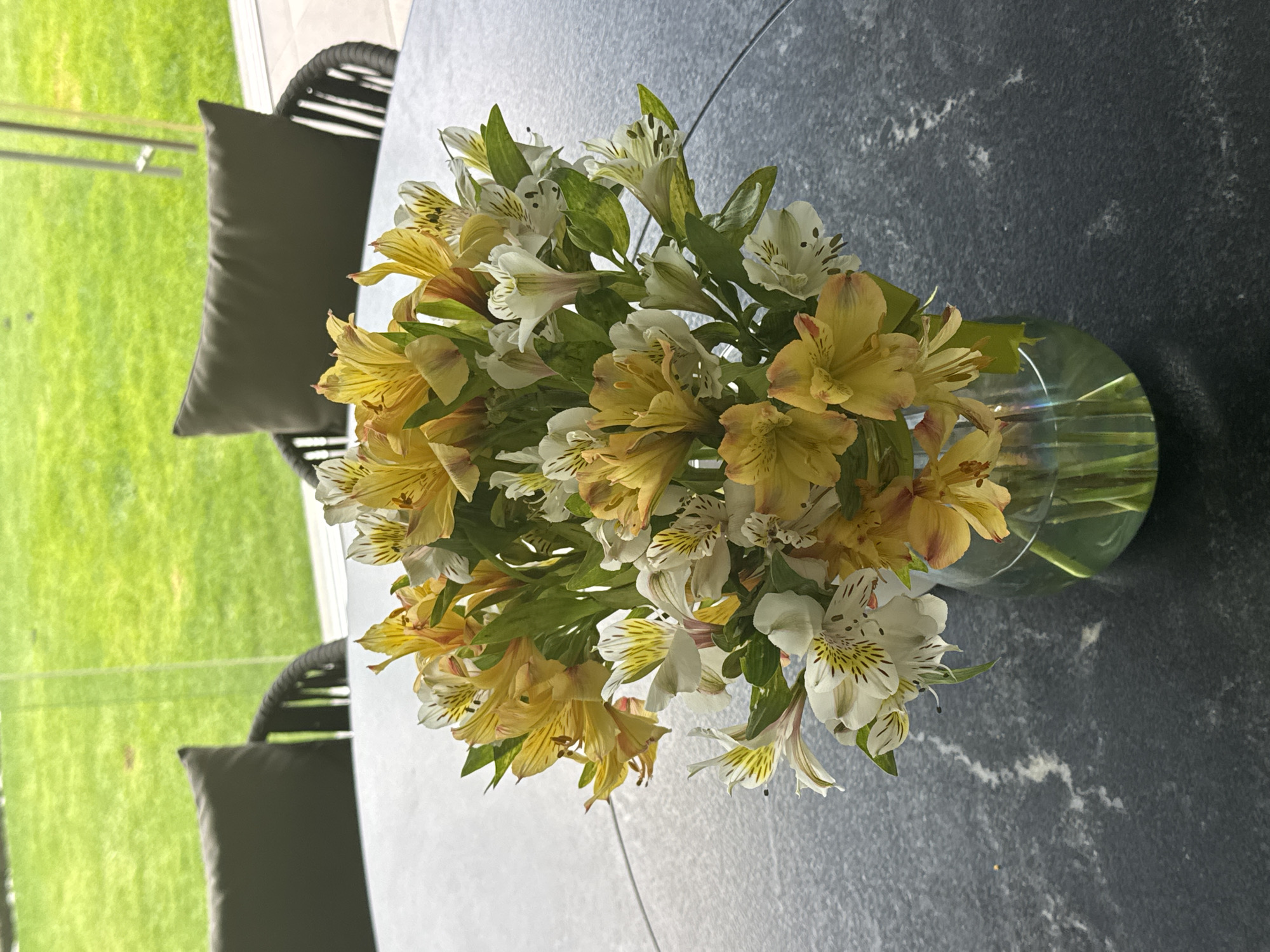 A high-angle shot captures a vase of flowers on a gray, textured, possibly stone, round table. The flowers are predominantly yellow and white alstroemeria lilies, mixed with green foliage. The vase is clear glass and appears to be partially filled with water.
Behind the table, one can see wicker chairs with gray cushions, set against a backdrop of green grass and a blurred view of what may be a window or glass partition.
Given the prompt includes a specific city, it is implied that this photo was taken in San Bernardino Tlaxcalancingo, Mexico. However, there are no visual cues within the image that independently verify the location.