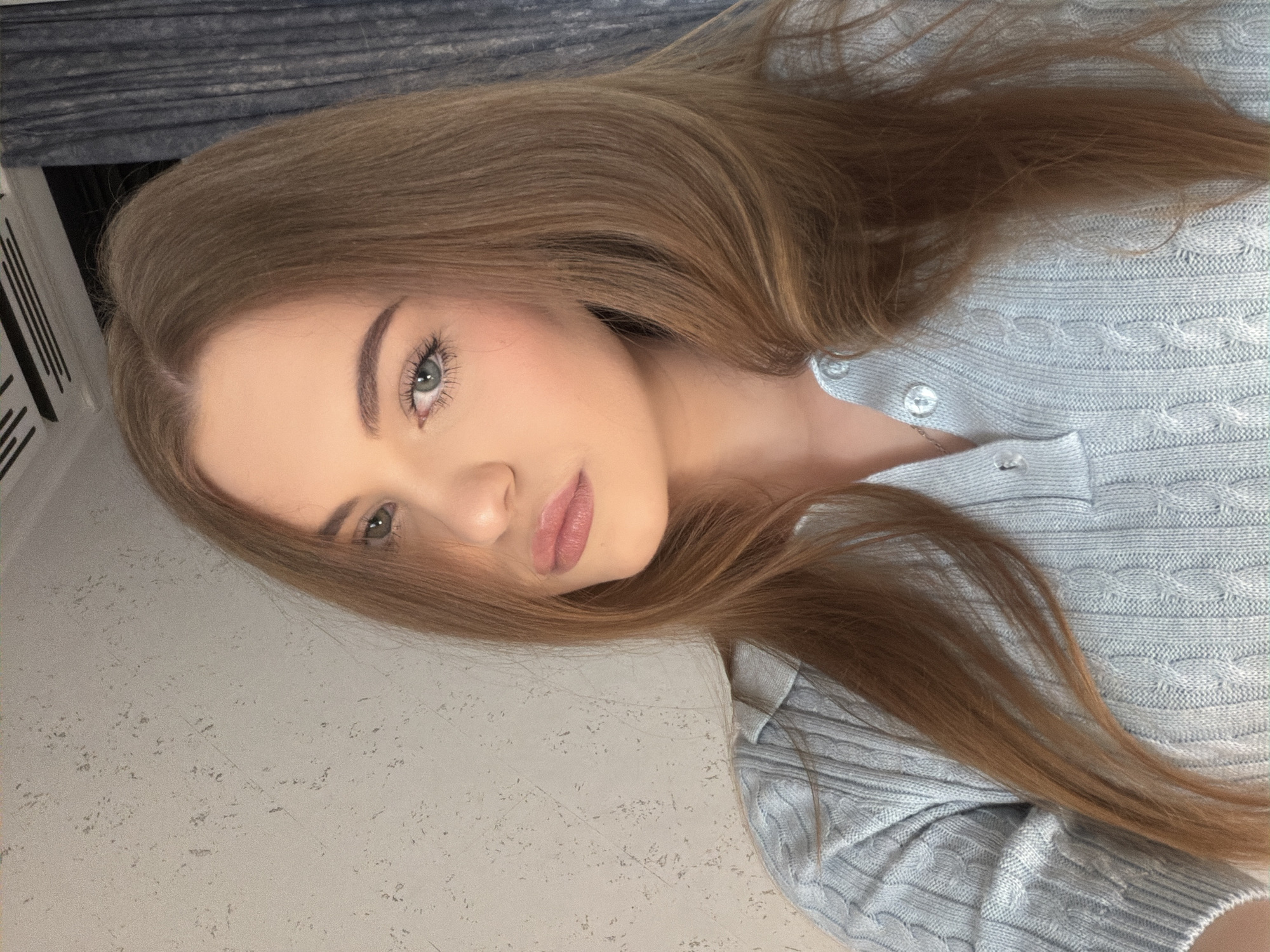 A young woman with shoulder-length auburn hair, green eyes, and fair skin is looking directly at the camera with a neutral expression. She is wearing a light blue cable-knit sweater with a collared neckline and two buttons. The background is a textured light gray wall with some dark blue curtains on the right side. The lighting appears to be natural, coming from the left. The image is a close-up, focusing on the woman's face and upper body. No specific location in Dnipro, Ukraine, can be determined from the image.