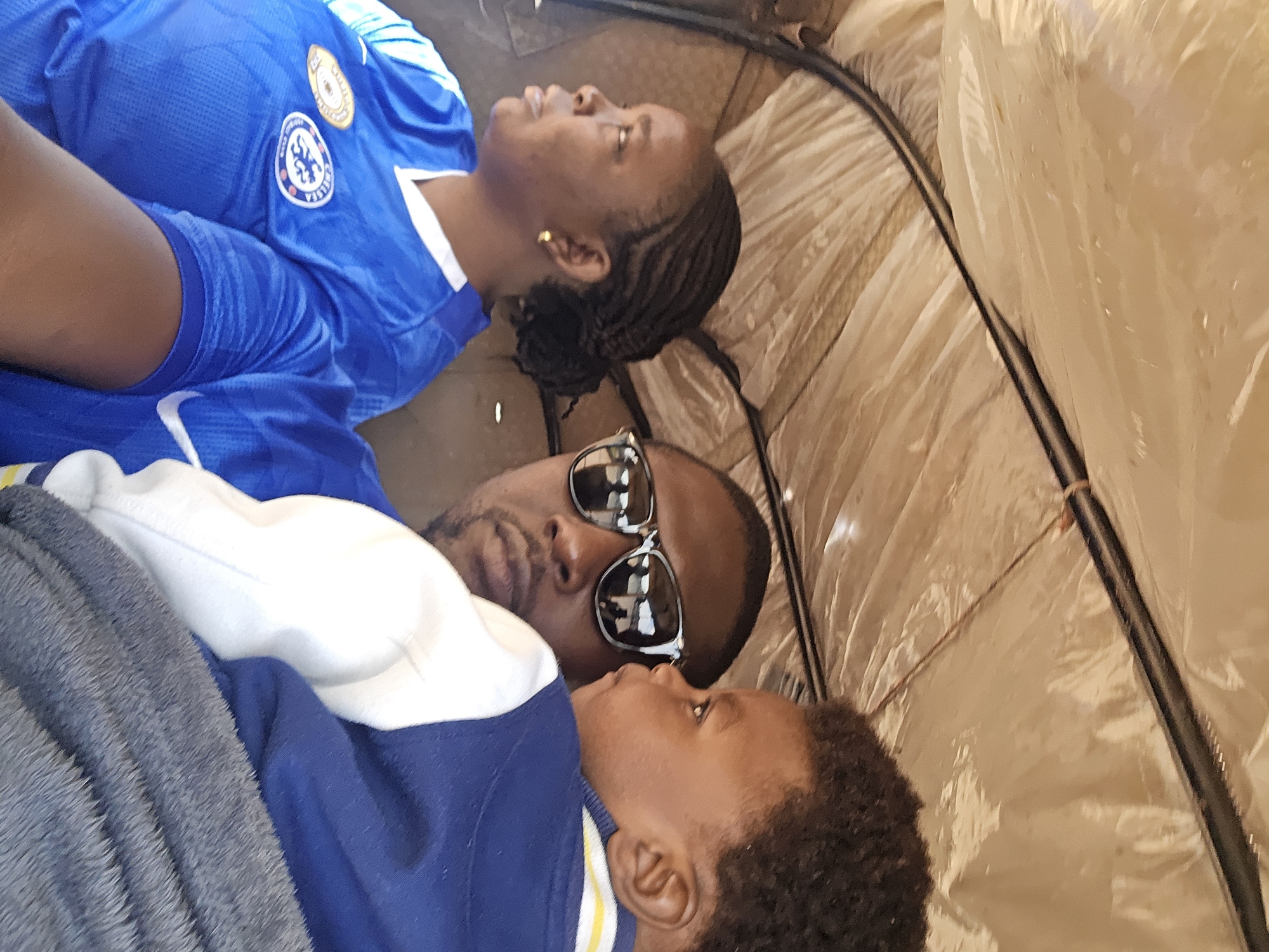 This is a close-up, indoor photograph taken from within a vehicle, likely a tuk-tuk, in Jos, Nigeria. The interior of the vehicle appears to be lined with a light brown, possibly plastic or tarp-like material, with visible support structures. Three individuals are the subjects of the image. On the left, a woman with braided hair is wearing a blue Chelsea Football Club jersey. In the center, a man with sunglasses is looking forward, holding a young child. The man is wearing a blue garment. The child, positioned on the man's right, is wearing a blue and white jacket. The lighting suggests it is daytime.
