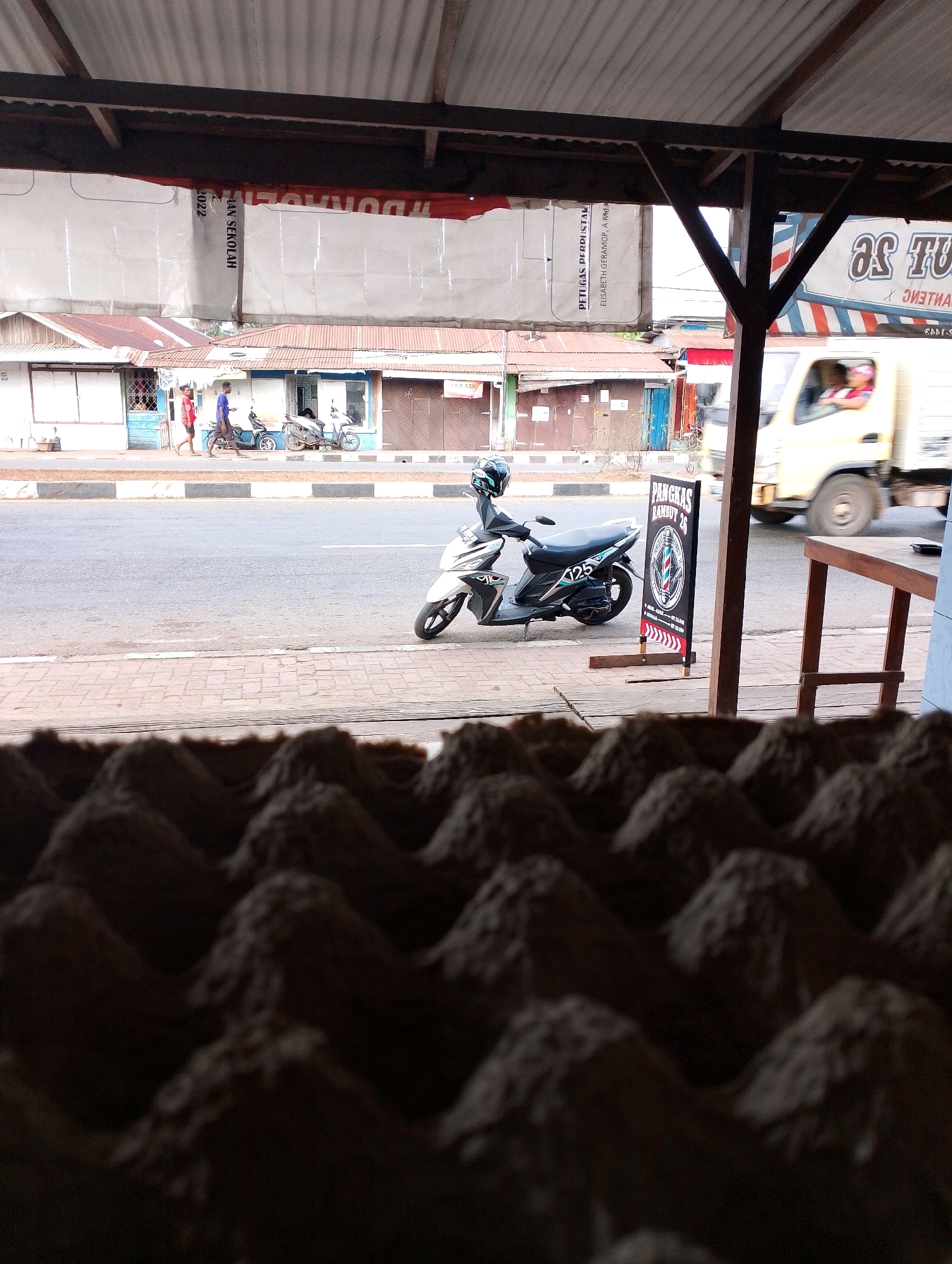 The image depicts a street scene in Merauke, Indonesia, viewed from under a corrugated metal awning. In the foreground, out of focus, is a textured surface resembling an egg carton. Beyond this, a paved road runs horizontally across the midground, with a sidewalk to its left. On the sidewalk, two young men walk towards the left, one in a red shirt and the other in a blue shirt, with a motorcycle parked between them. Further to the left, more motorcycles are visible outside a row of buildings with tin roofs, suggesting a commercial area. A white and black scooter is parked on the sidewalk to the right of the road, featuring a "125" decal and a helmet on the handlebars. Next to the scooter is a freestanding sign for "Pangkas Rambut 26," a barbershop, displaying a barber pole emblem. In the background, a yellow truck is moving from left to right on the road, its driver visible behind the windshield. A blurred sign with text in Indonesian, possibly advertisements or announcements, is stretched across the upper part of the scene. The overall environment suggests a typical roadside commercial strip in an Indonesian city.
