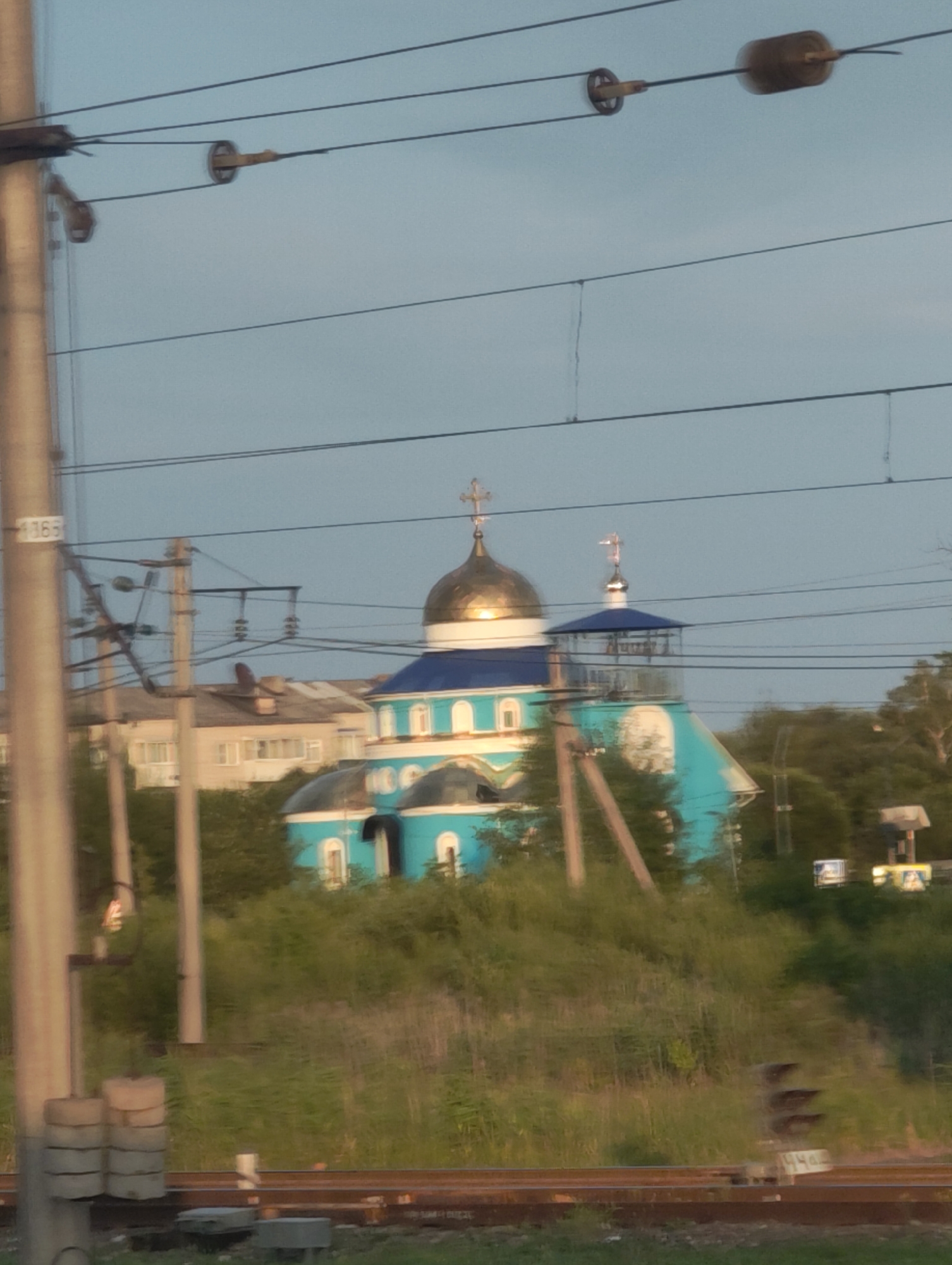In Smidovich, Russia, a clear sky is visible above a turquoise church with golden domes, featuring a prominent bell tower and multiple crosses. The church is situated on a grassy incline, partially obscured by trees. In the foreground, train tracks run horizontally across the image, with a railway signal visible on the right. Power lines crisscross the sky, supported by utility poles. A few buildings are visible behind the church, and a portion of a larger pole with insulators stands to the left. The overall scene depicts a serene, possibly late afternoon, view of a religious building near a railway line in a Russian town.