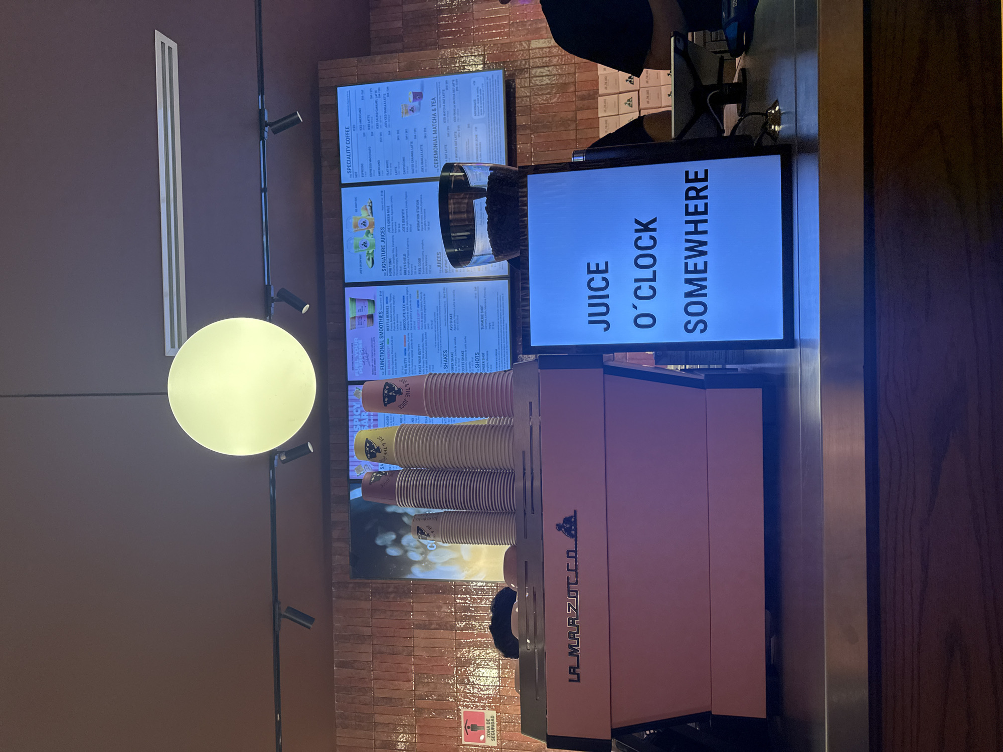 This image depicts the interior of a Joe & The Juice location, likely a franchise, showcasing a service area and various elements related to the business.
The view appears to be taken from behind the service counter, providing a perspective of the customer-facing side. A large, pink La Marzocco espresso machine dominates the foreground, with stacks of disposable cups placed above it. Above the espresso machine is a display showing the store's menu which includes various beverages such as specialty coffee, signature juices, and ceremonial matcha. A display board with the text "JUICE O'CLOCK SOMEWHERE" can be seen in front of the machine.
The environment consists of tiled walls and warm lighting with a large globe light above the counter area. In the upper part of the shot, a dark-clad person stands behind the counter, likely a staff member, partially visible at the edge of the frame.
The location of the Joe & The Juice is assumed to be in Mexico City, Mexico.
