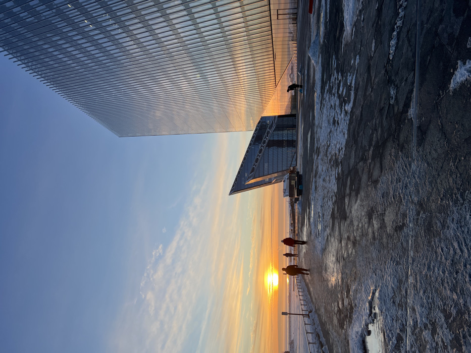 The image depicts a winter scene at the Lakhta Center complex in Saint Petersburg, Russia, during sunset or sunrise. The prominent subject is the Lakhta Center skyscraper, a tall, multi-faceted glass structure, dominating the right side of the frame. Its reflective surface captures the vibrant orange and yellow hues of the low sun. To its left, a smaller, angular glass building, also part of the complex, is visible.
The foreground consists of a dark paved plaza and a pathway, both intermittently covered with patches of snow and ice. On the pathway, along the left side of the frame, three individuals are walking away from the viewer, appearing as silhouettes against the bright horizon. A dark metal railing runs parallel to the path. On the plaza, near the base of the Lakhta Center skyscraper, a fourth individual is observed bent over, engaged in clearing snow or debris with a tool.
The sky transitions from a clear light blue at the top to intense orange and yellow above the horizon, with thin, elongated clouds. The sun is a distinct, bright orb positioned just above the horizon line over what appears to be a body of water, likely the Gulf of Finland, which exhibits icy or partially frozen conditions.