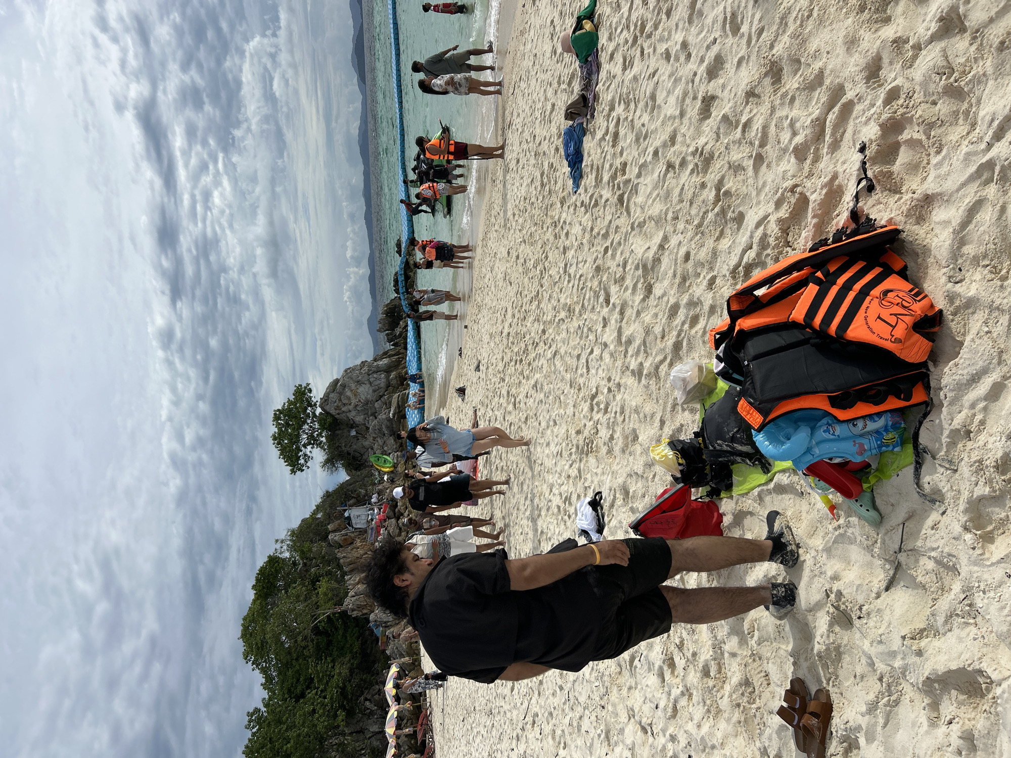 In Tambon Phru Nai, Thailand, on a crowded beach with white sand, a group of people are visible. The sky is overcast with gray clouds. In the foreground, a man in a black short-sleeved shirt and black shorts stands facing away from the camera, looking down. Near his feet, brown sandals and a pile of beach gear, including orange and black life vests, bags, and a blue inflatable toy, are scattered on the sand. Further down the beach, individuals are gathered near the turquoise water, some wading in or standing by inflatable flotation devices. In the background, rocky outcrops with vegetation rise from the sea, and a line of blue floating barriers separates the swimming area. Several colorful umbrellas are set up on the sand further inland.