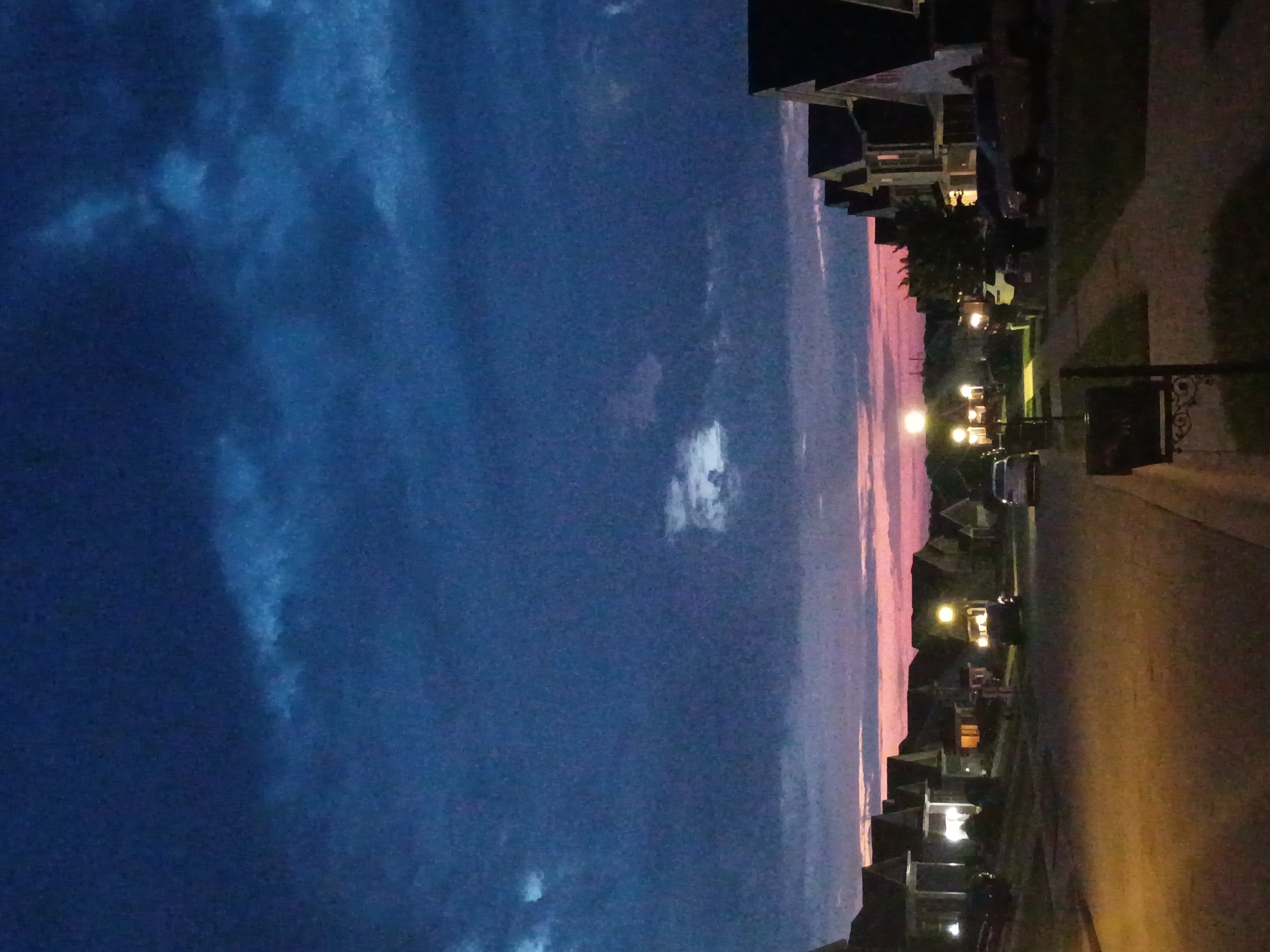 The image depicts a suburban street at dusk. The sky is a deep, dark blue with scattered clouds, and a hint of pink and orange is visible on the horizon, indicating the last light of sunset. The street is paved and appears to be a residential area with houses lining both sides. The houses are of modern suburban architecture with pitched roofs and illuminated windows, suggesting that some residents are home or that exterior lights are on. Several vehicles are parked on the street and in driveways, and streetlights cast a warm glow on the scene. There are no people visible in the image. The overall atmosphere is one of quiet twilight in a residential neighborhood. The image does not contain sufficient information to determine the specific location within the city of Richmond, United States of America.
