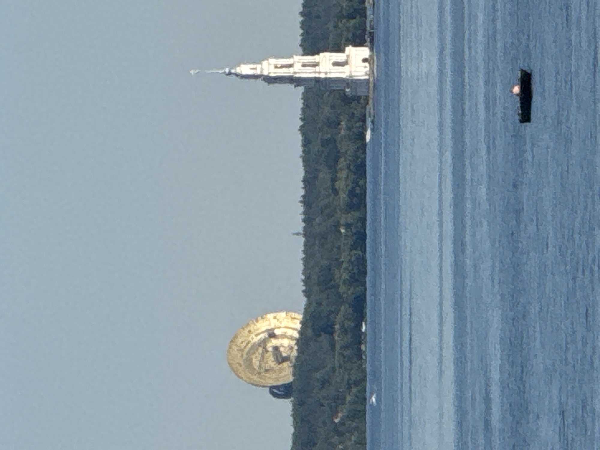 This image captures a wide view of the Volga River in Volzhanka, Russia, on a clear, sunny day. In the distance, silhouetted against the pale blue sky, is a large, golden, disc-shaped structure partially obscured by a dense green forest. To the right of the golden structure, also on the shoreline, stands a tall, white, multi-tiered bell tower, topped with a spire. The river itself is a calm expanse of blue water, reflecting the sky. In the foreground, a small, dark boat with a glowing pink object is visible on the water. The scene suggests a tranquil, scenic landscape with distinct architectural landmarks.