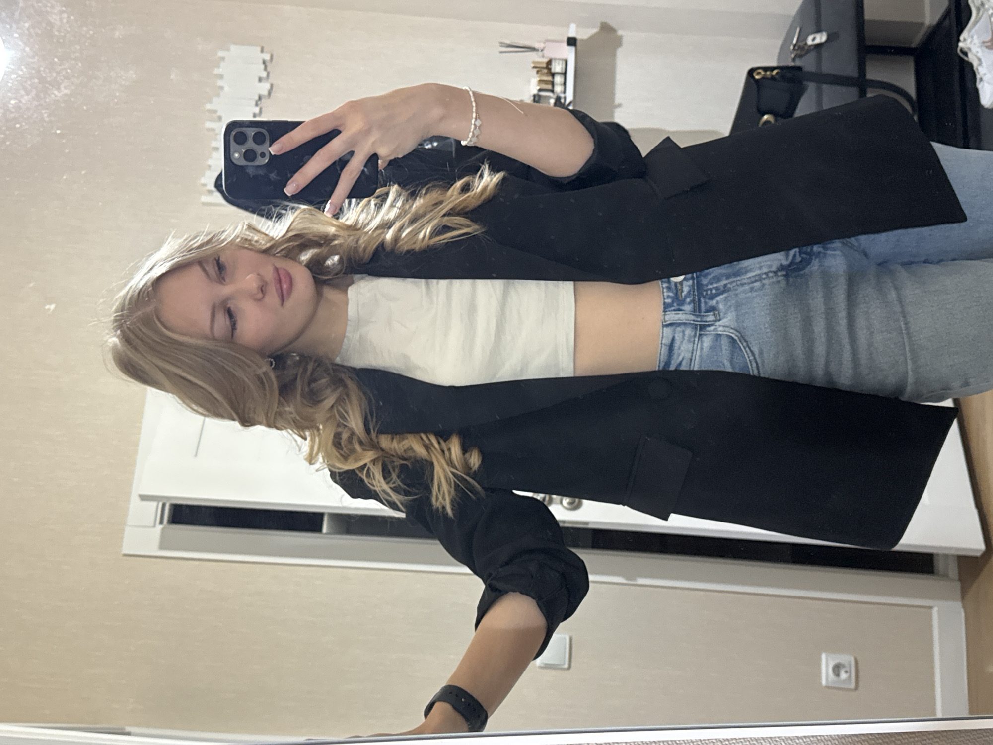 A young blonde woman is taking a selfie in a mirror. She is wearing a black blazer over a white crop top and light-wash jeans. She has curly blonde hair and is wearing minimal jewelry. The background appears to be a room, possibly a bedroom or dressing area, with a door and a light switch visible. There is a shelf with bottles and a purse on a stool in the background. The overall scene suggests a casual indoor setting. The image does not contain any visual cues to pinpoint a specific city like Perm, Russia.