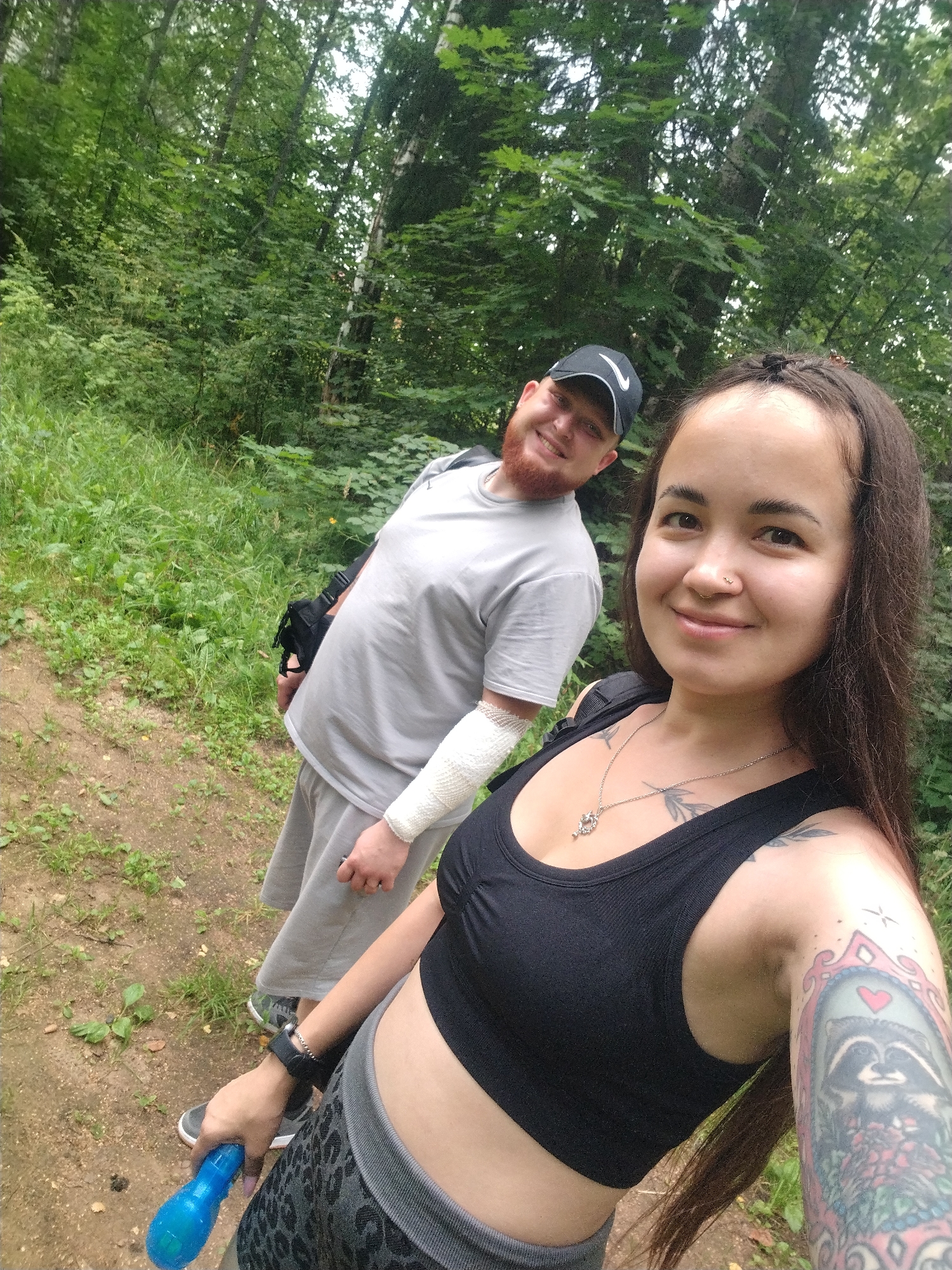 Two people, a man and a woman, are posing for a selfie in a wooded area. The man, on the left, is wearing a grey t-shirt, grey shorts, and a black baseball cap. He has a beard and is smiling at the camera. His right arm is wrapped in a white bandage. The woman, on the right, is wearing a black crop top and grey patterned leggings. She has long dark hair, a nose ring, and tattoos on her arm. She is holding a blue object in her right hand and is smiling. They are standing on a dirt path with green foliage and trees in the background. The location appears to be a park or forest. The weather is overcast.