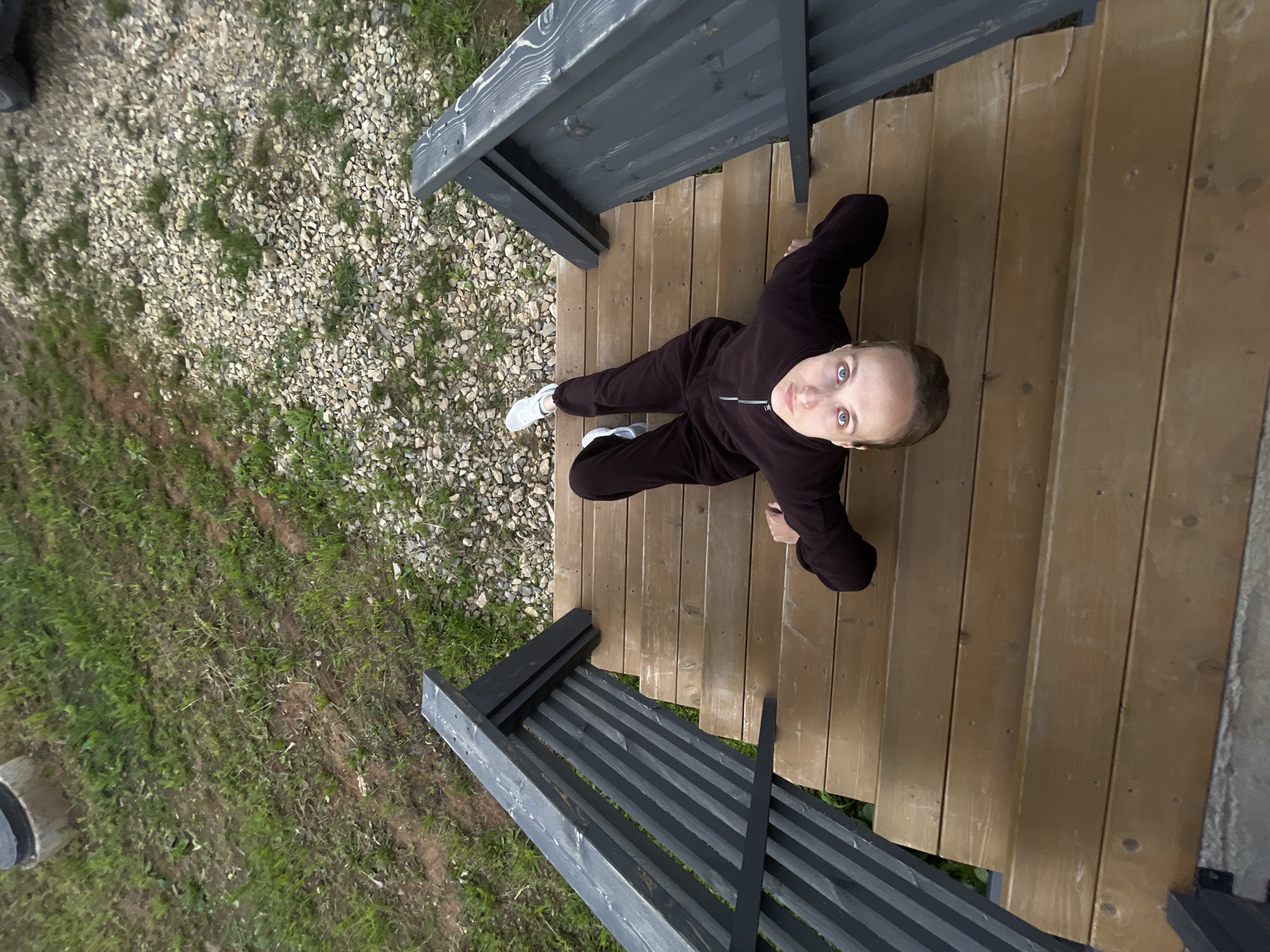A person is sitting on wooden stairs, looking up at the camera. The person is wearing a dark tracksuit and white sneakers. The stairs are outdoors, with gravel and grass visible to the sides. There are dark grey wooden railings on either side of the stairs. The scene appears to be in a rural or suburban setting. The lighting suggests it might be daytime, possibly overcast. The person's expression is neutral. No text is visible in the image.