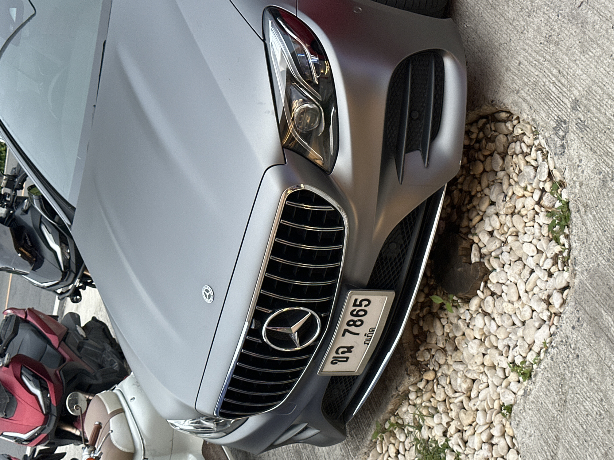 The image shows a close-up of the front grille and headlights of a matte gray Mercedes-Benz E-Class sedan. A license plate with Thai characters and the number "7865" is mounted below the grille. In the background, two motorcycles are parked, one red and one black. The car is parked on a textured concrete surface, with a patch of small white stones to the left of the front bumper. The scene is captured during daylight. The location is identified as Karon, Thailand.
