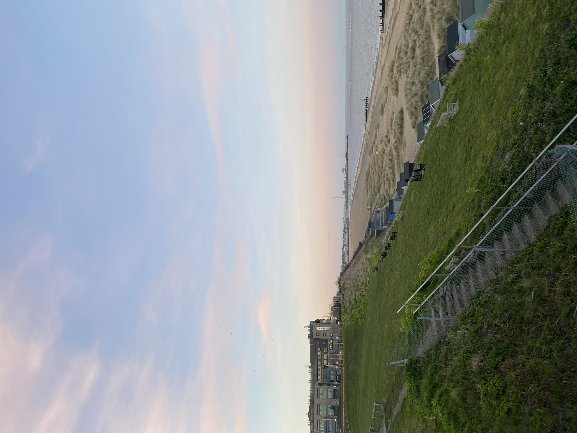 The image depicts a coastal scene in East Suffolk, United Kingdom. The sky is a pale gradient of blue and pink, suggesting either sunrise or sunset. Below the sky is the sea, followed by a sandy beach. Along the beach are rows of beach huts. Adjacent to the beach is a grassy area, descending towards a building complex. A set of concrete steps with a metal railing runs down the slope. The overall scene captures a calm, coastal environment.