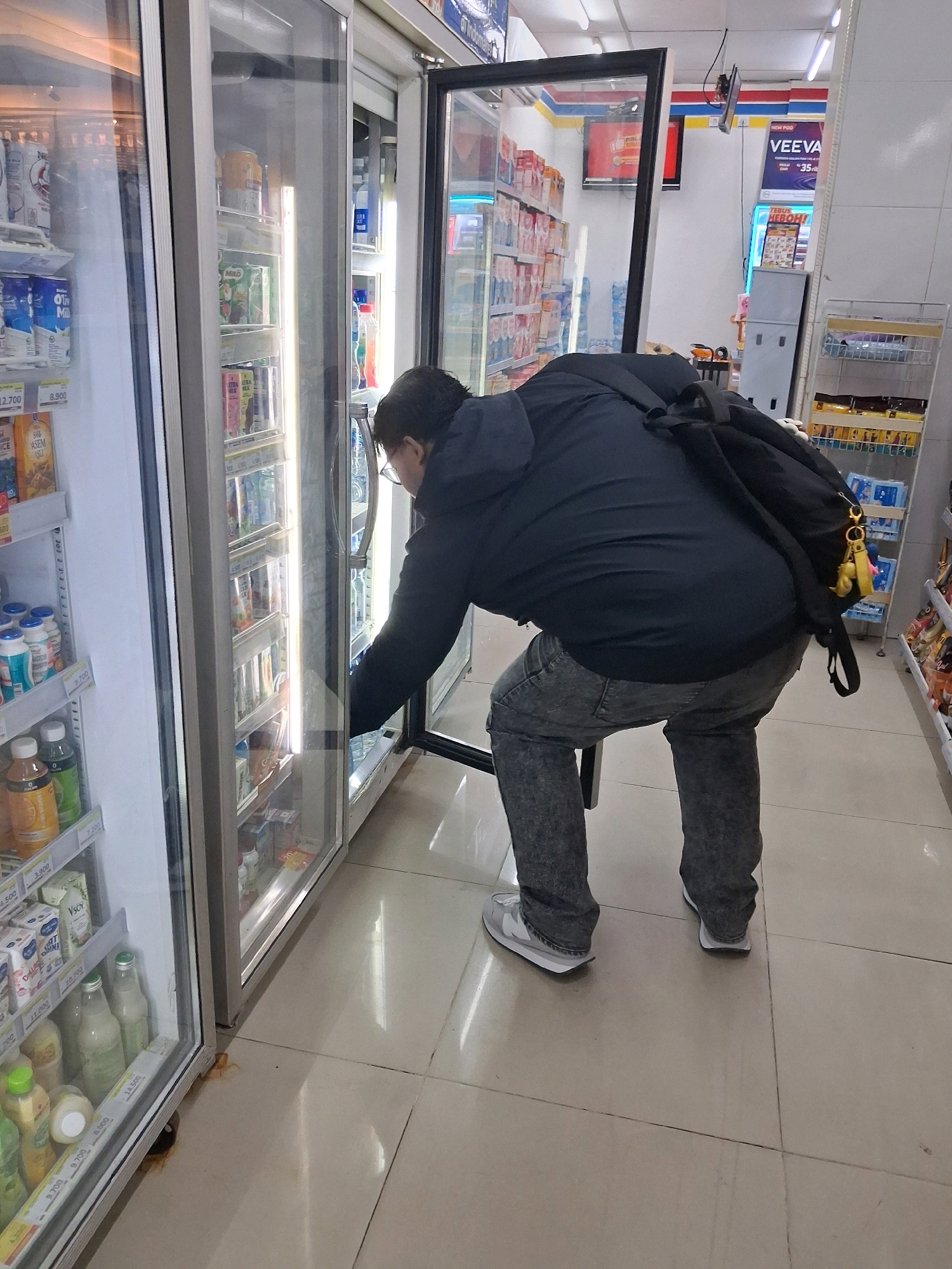 The image depicts a person in a convenience store in Surakarta, Indonesia. The individual, wearing a dark jacket and grey jeans, is bent over and reaching into a refrigerated display case filled with beverages. A black backpack is slung over their shoulders, with a yellow keychain attached. The store's interior features tiled flooring and bright artificial lighting. Various shelves stocked with products are visible in the background. The refrigerator doors are glass, allowing a clear view of the items inside, including cans and bottles of drinks. The overall scene captures a mundane moment of shopping within a retail environment.
