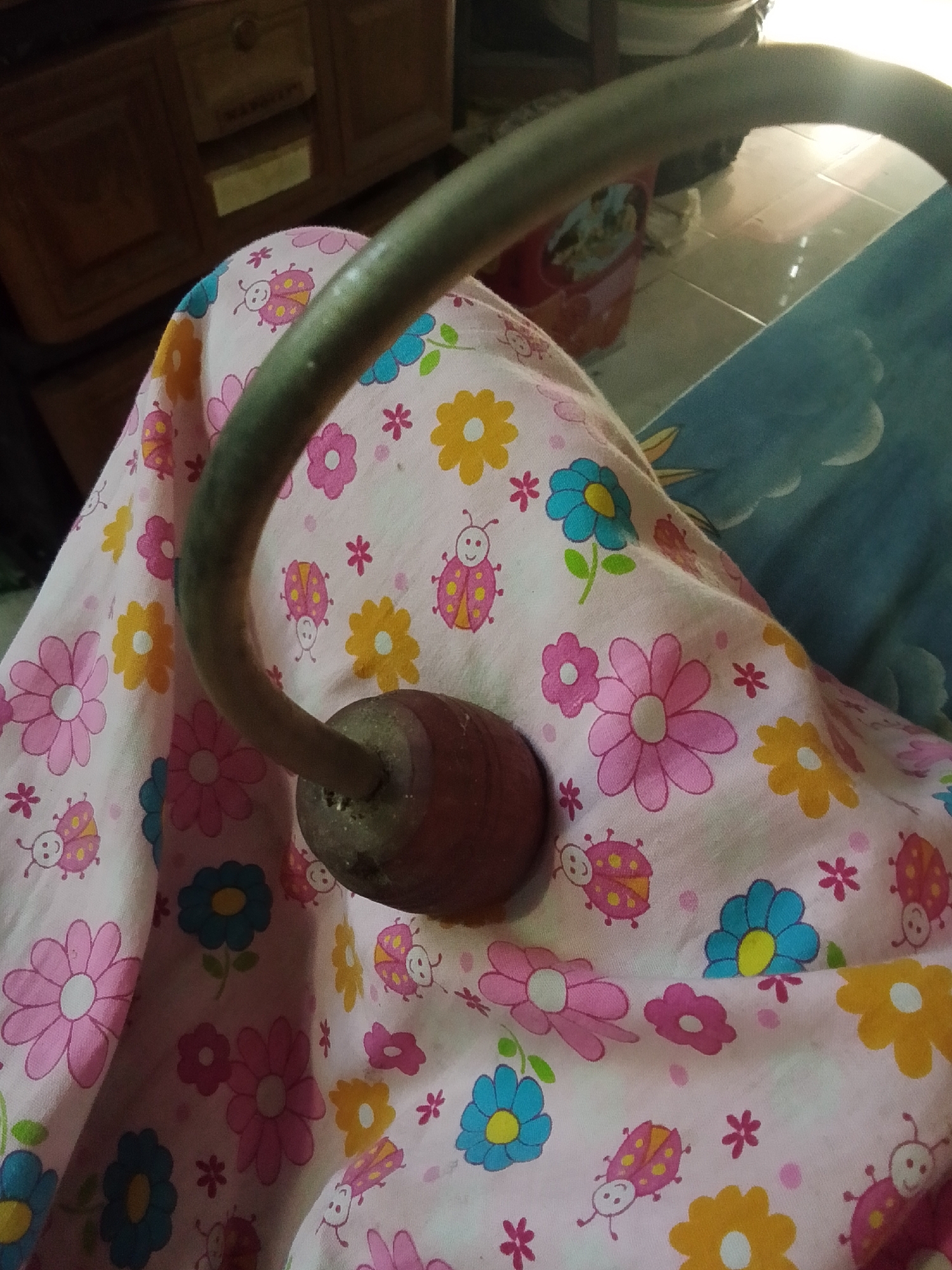 The image shows a close-up view of a person's lap covered with a pink fabric patterned with flowers and ladybugs. A curved metal object with a wooden cylindrical end is resting on the fabric. The background is blurred, showing a wooden cabinet and a tiled floor. The scene is indoors, possibly in a residential setting in Bulurejo, Indonesia.