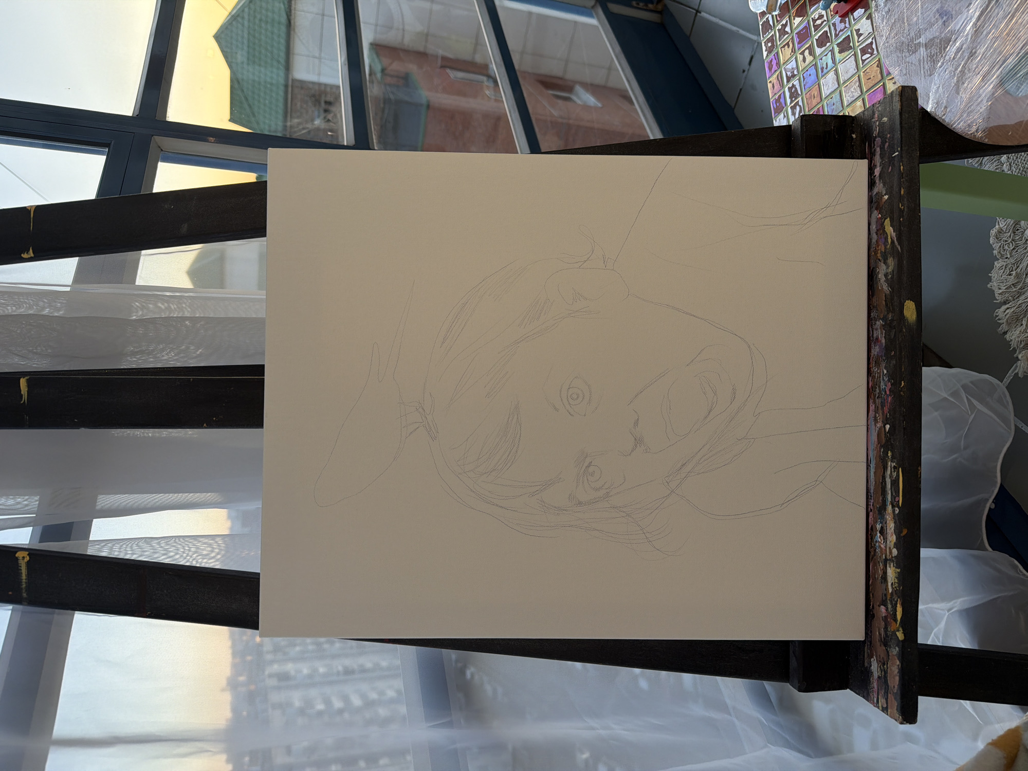 This media file depicts a pencil sketch of a person's face, angled downwards, on a white canvas. The sketch is in progress, with unfinished lines defining the hair, eyes, nose, and mouth. The canvas is set up on a dark wooden easel, which shows visible paint smudges, suggesting it is being used for art creation.
The scene is set indoors, likely an art studio or a room with a large window. Through the window, reflections of buildings and the sky are visible, hinting at an urban environment. The lighting suggests it might be daytime, possibly late afternoon or early morning, given the warm, golden hue of the light reflecting in the window. The overall atmosphere appears calm and focused, centered on the artistic endeavor. No visible text is present in the image. The location is identified as Krasnoyarsk, Russia.