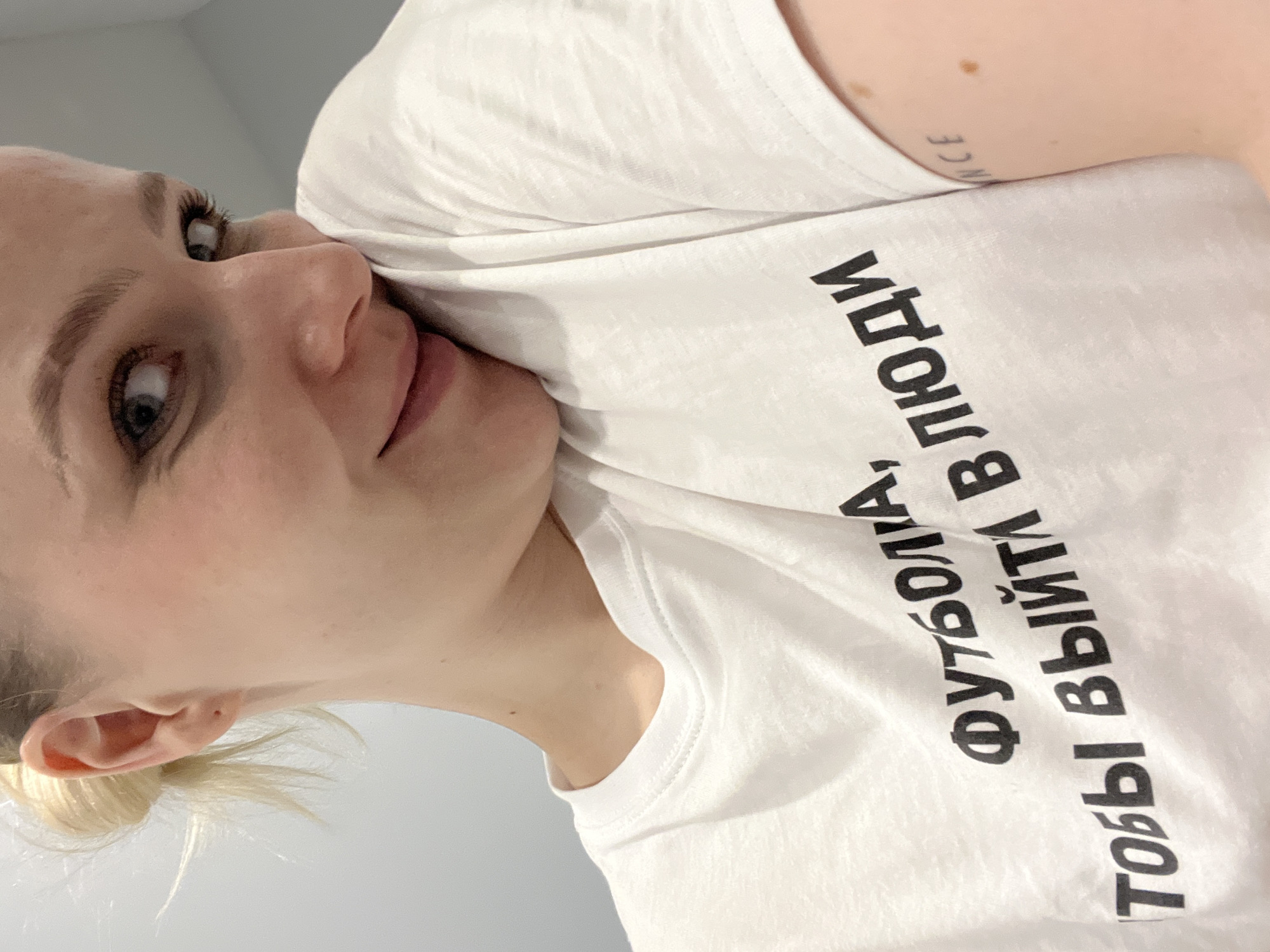 A head-and-shoulders close-up shot depicts a woman with fair skin and light hair. She is wearing a white T-shirt with black text in Russian, which reads "ФУТБОЛА, ТОБЫ ВВЫЙТИ В ЛЮДИ". The shirt obscures the lower portion of her face, slightly pushing her cheek up. Her expression appears pensive, and she is looking towards the upper left of the frame. A small portion of a tattoo, featuring the word "ENCE" is visible on her upper arm. The background is a plain, light-colored wall. No details indicate this photograph was taken in the city of Khimki, Russia.