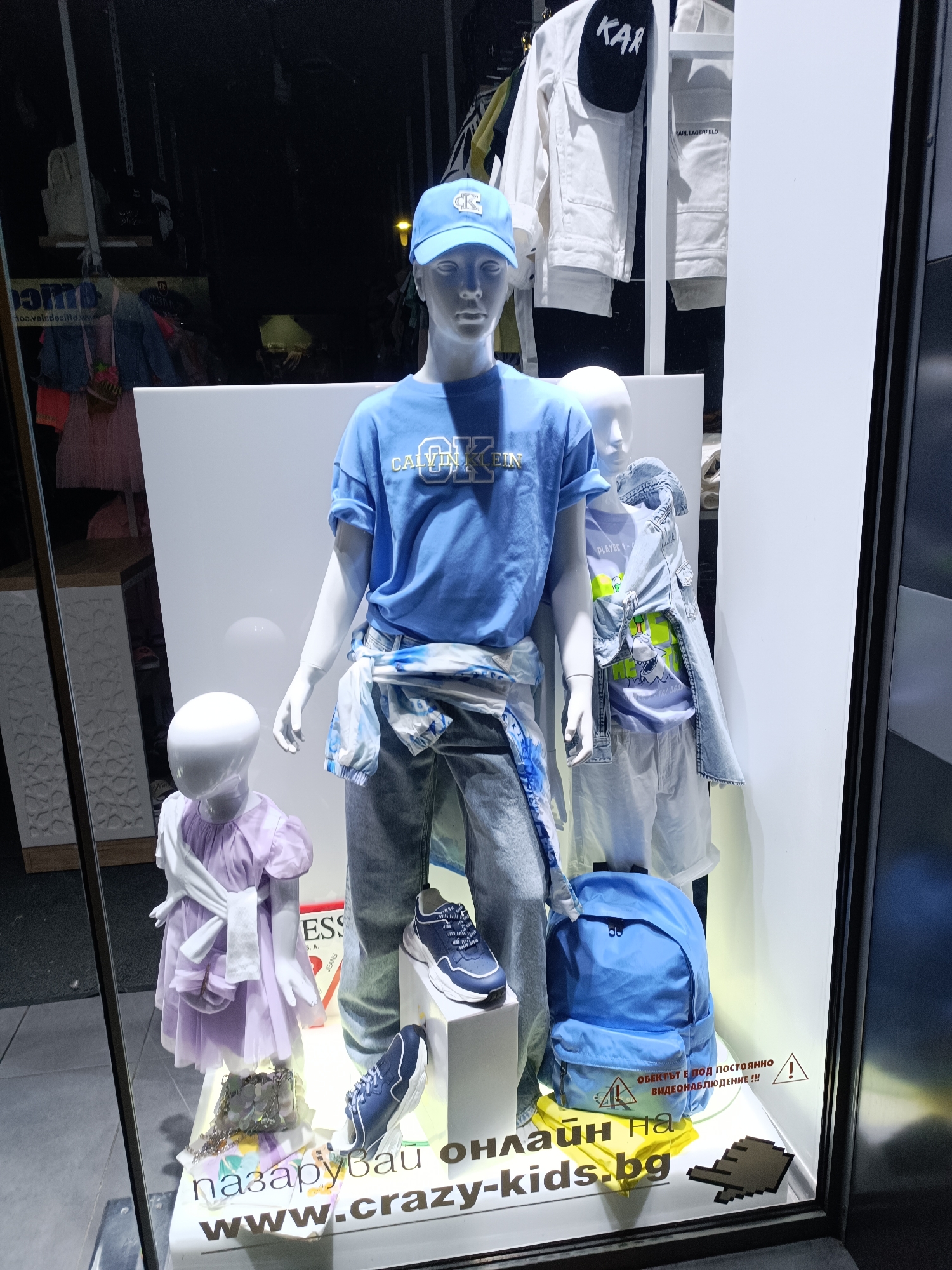 This is a nighttime photograph of a children's clothing store window display in Pleven, Bulgaria. The brightly lit display features three mannequins showcasing various outfits and accessories, visible through the reflective glass.
The central and tallest mannequin, representing an older child or teenager, wears a light blue short-sleeved t-shirt with "CALVIN KLEIN" in white lettering across the chest. It is paired with light-wash jeans and blue sneakers with chunky white soles. A light blue and white tie-dye shirt is tied around its waist, and it wears a light blue baseball cap with a white logo. A large, plain blue backpack sits on the floor beside it.
To the right, a slightly smaller mannequin displays a light purple t-shirt featuring a green graphic print of a shark and the partial text "PLAYER 1-". It wears white shorts, and a light blue denim jacket is draped over its shoulders.
On the left, a small child-sized mannequin is dressed in a light purple, short-sleeved dress with a white sash tied around its waist. A small, shiny silver sequined handbag rests on the display base in front of it.
In the background, behind the mannequins, a white denim jacket with "KARL LAGERFELD" printed on it and a black cap with "KAR" are visible. Another small sign featuring "GUESS JEANS" is present near the child mannequin.
The base of the display includes text in Bulgarian: "пазарувай онлайн на www.crazy-kids.bg" (shop online at www.crazy-kids.bg), accompanied by a pixelated hand cursor icon. A prominent warning sticker reads "ОБЕКТЪТ Е ПОД ПОСТОЯННО ВИДЕОНАБЛЮДЕНИЕ !!!" (The object is under constant video surveillance!!!), with an associated exclamation mark icon. Reflections on the window glass include blurred lights and a reversed "100.velsdeilto w".