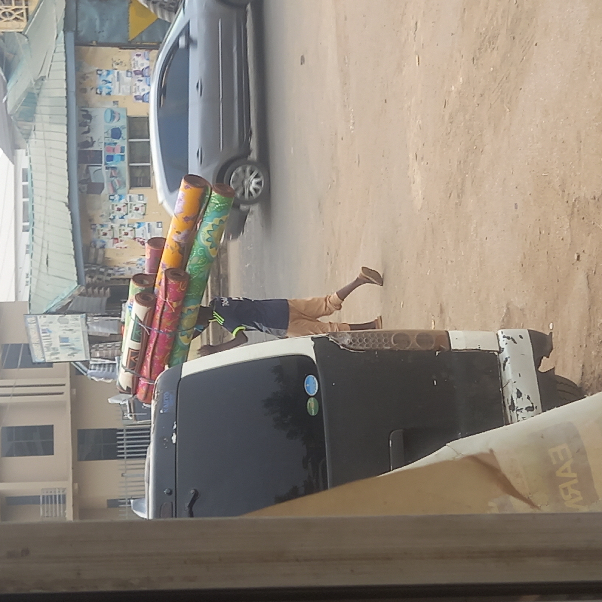 The image, taken in Dadi Riba, Nigeria, shows a street scene rotated 90 degrees clockwise. In the foreground, a person is pushing a heavily loaded three-wheeled utility vehicle or cart. The person, appearing to be a young male, wears a dark blue t-shirt with lime green accents and light brown trousers. He has dark skin and is walking barefoot or in open sandals.
The utility vehicle, which has a black cabin and a white front section, is laden with numerous brightly colored rolls, likely rugs, mats, or textiles. These rolls are secured with ropes and display various patterns in yellow, green, pink, and orange. Small circular decals, one blue and one green/yellow, are visible on the vehicle's black body.
In the background, a grey car is visible on what appears to be an unpaved, sandy road. Several buildings line the street; one has a light-colored facade with rectangular windows, and another, further back, features a corrugated metal roof and various posters or signs on its walls. A large, unreadable sign with text and images is also present on the left side.
The scene depicts daily life and commerce, with the individual transporting goods under bright daylight, suggesting sunny and clear weather. The architecture and street conditions are characteristic of a commercial area in a developing urban or semi-urban region. Mirrored letters that appear to spell "ЕЯАЯ" are visible on a blurred foreground object, likely a window or frame through which the photo was taken.