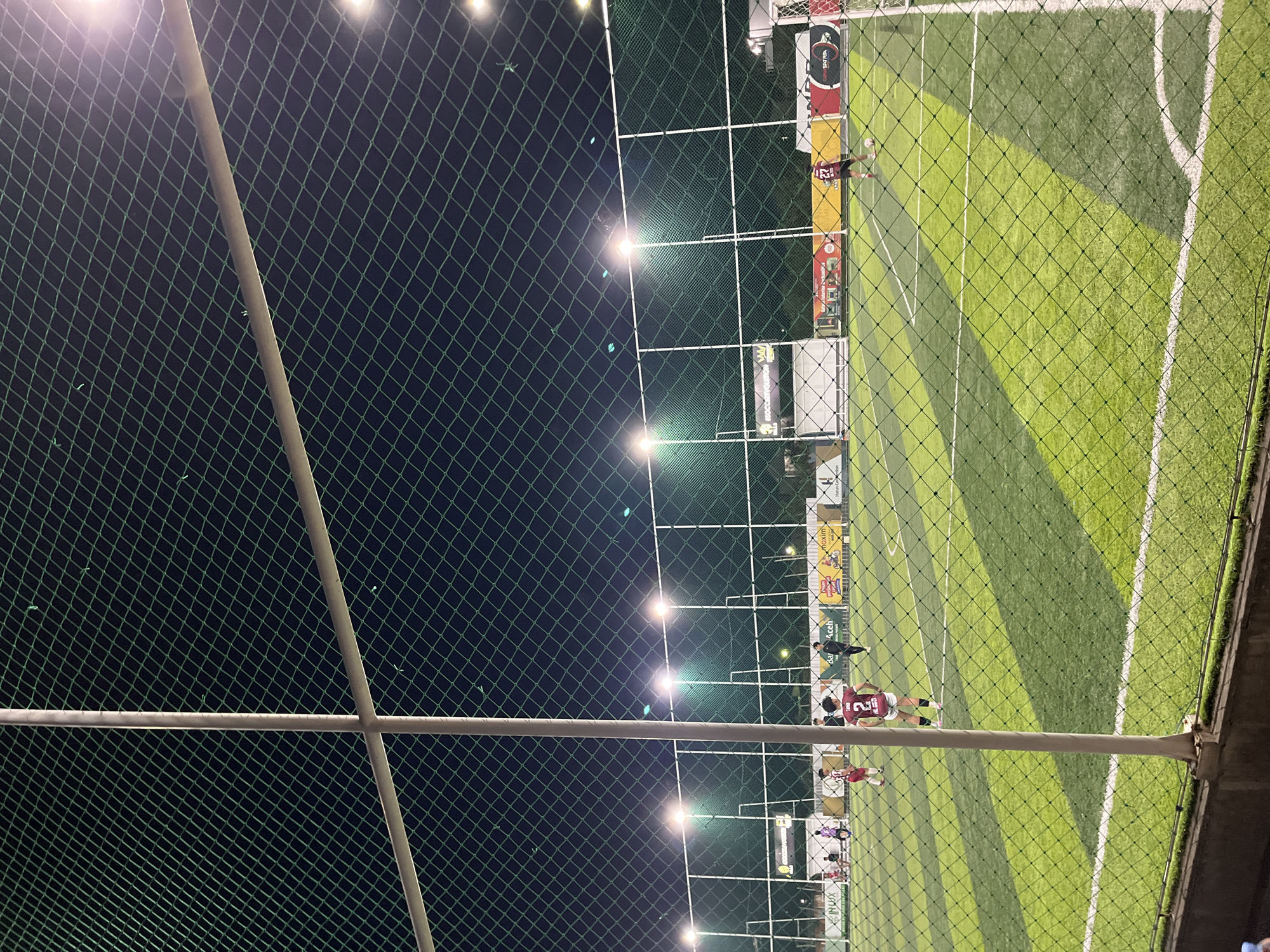 The image shows an artificial soccer field at night, taken through a green net and support poles. The field is illuminated by overhead lights, and a few people are visible playing soccer, wearing jerseys with numbers, including one visible number "21". Advertising boards line the side of the field. The location is identified as being in Lhokseumawe, Indonesia.