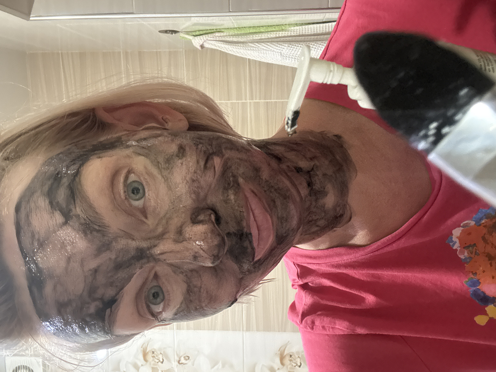 A woman with blonde hair and blue eyes, her face covered in a black charcoal mask, is looking at the camera. She is wearing a pink t-shirt. A black makeup brush is in the foreground, out of focus, with a white pump dispenser behind it. The background appears to be a bathroom with tiled walls and a towel hanging on a hook. The lighting is bright, suggesting it is daytime. The image is a selfie taken indoors. There is no discernible information to place the location in Sochi, Russia.