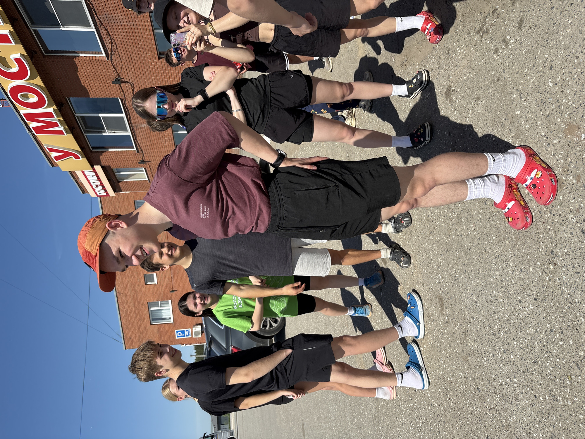 A group of approximately eight young people stands in a line on a paved street in front of a brick building with a yellow sign that reads "MOCKBA" vertically. The sky is clear and blue.
The individuals are dressed in casual clothing, including shorts, t-shirts, and sneakers or Crocs. Some are wearing hats or sunglasses. They appear to be posing for a photo or video, as one person at the top of the frame is holding a phone and taking a picture.
In the background, a dark colored car is visible, along with a "P" parking sign.