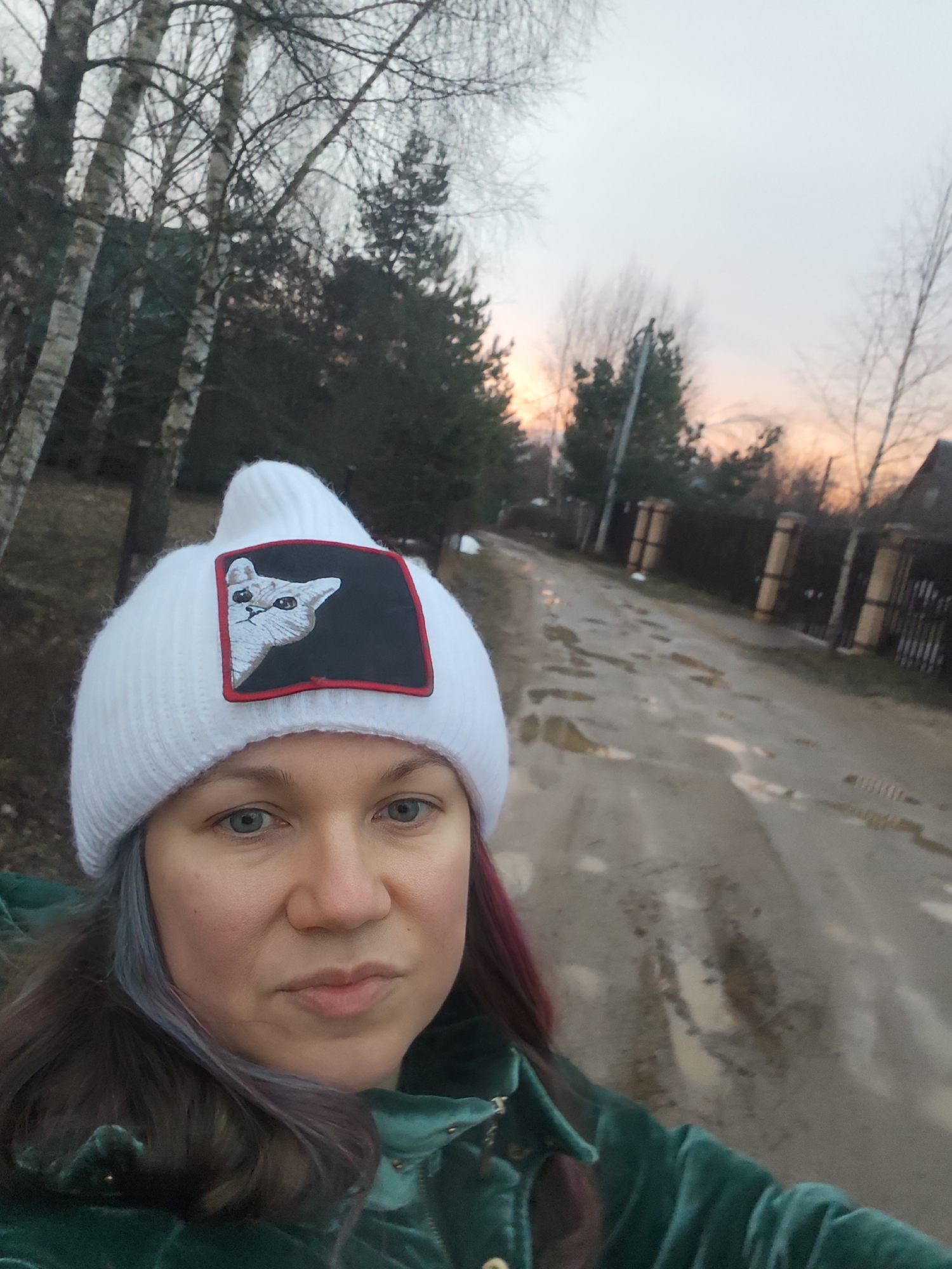 A person wearing a white knit hat with a cat patch and a green velvet jacket is taking a selfie. The background shows a muddy road with puddles, a fence, and trees without leaves. The sky is a pale orange, indicating sunset or sunrise. The location is Свистуха (кузяевский С/о), Russia.