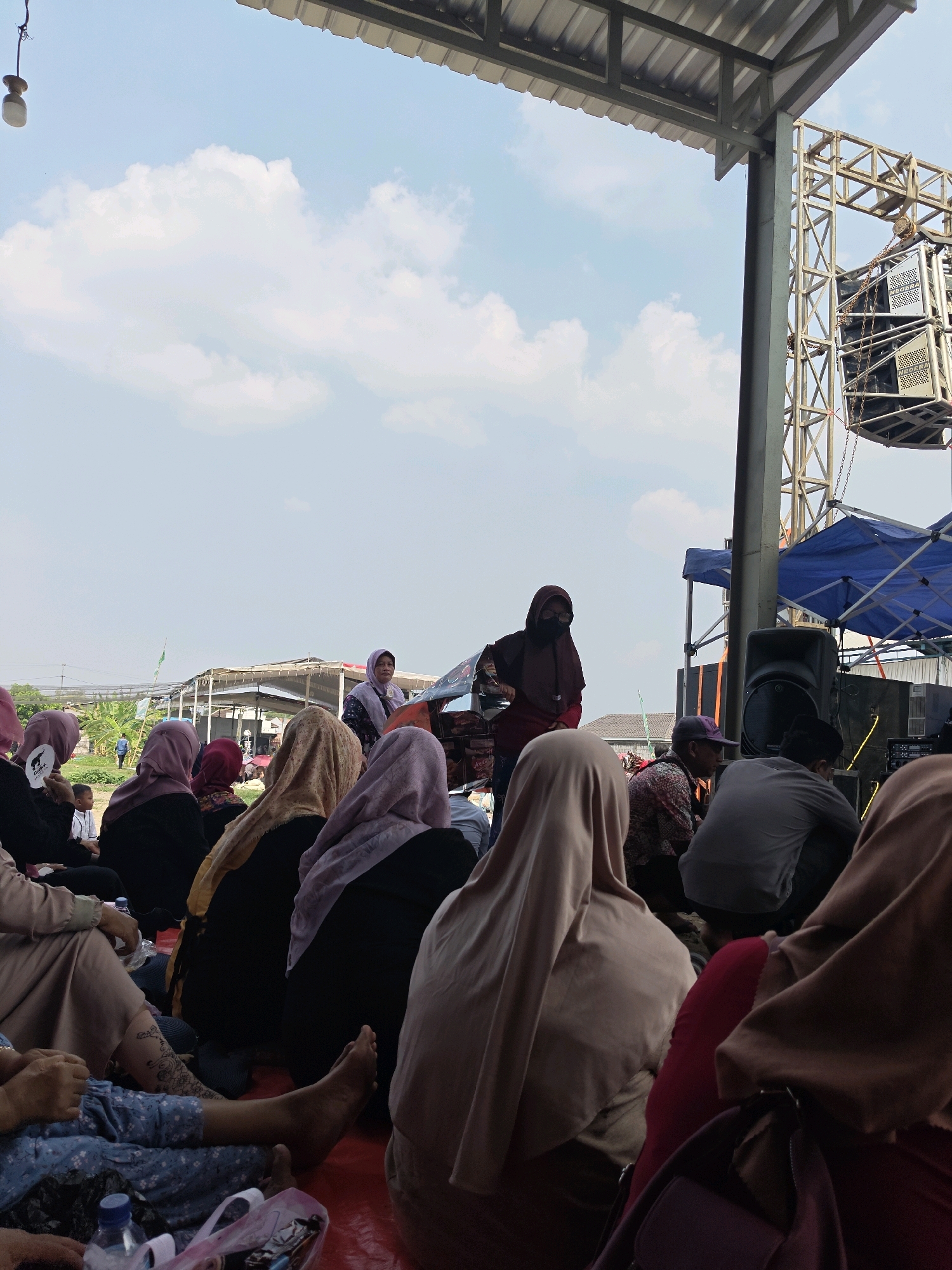 This image depicts an outdoor gathering of people, likely in Bangsal, Indonesia, under a covered structure. A group of individuals, predominantly women wearing headscarves, are seated on the ground, facing a platform. On the platform, two women are standing, one of whom appears to be speaking or addressing the seated audience. Behind them, a large speaker system is mounted on a metal truss structure, suggesting a performance or event is taking place. The environment appears to be open, with a bright sky and some vegetation visible in the background. The overall scene suggests a community event or religious gathering.
