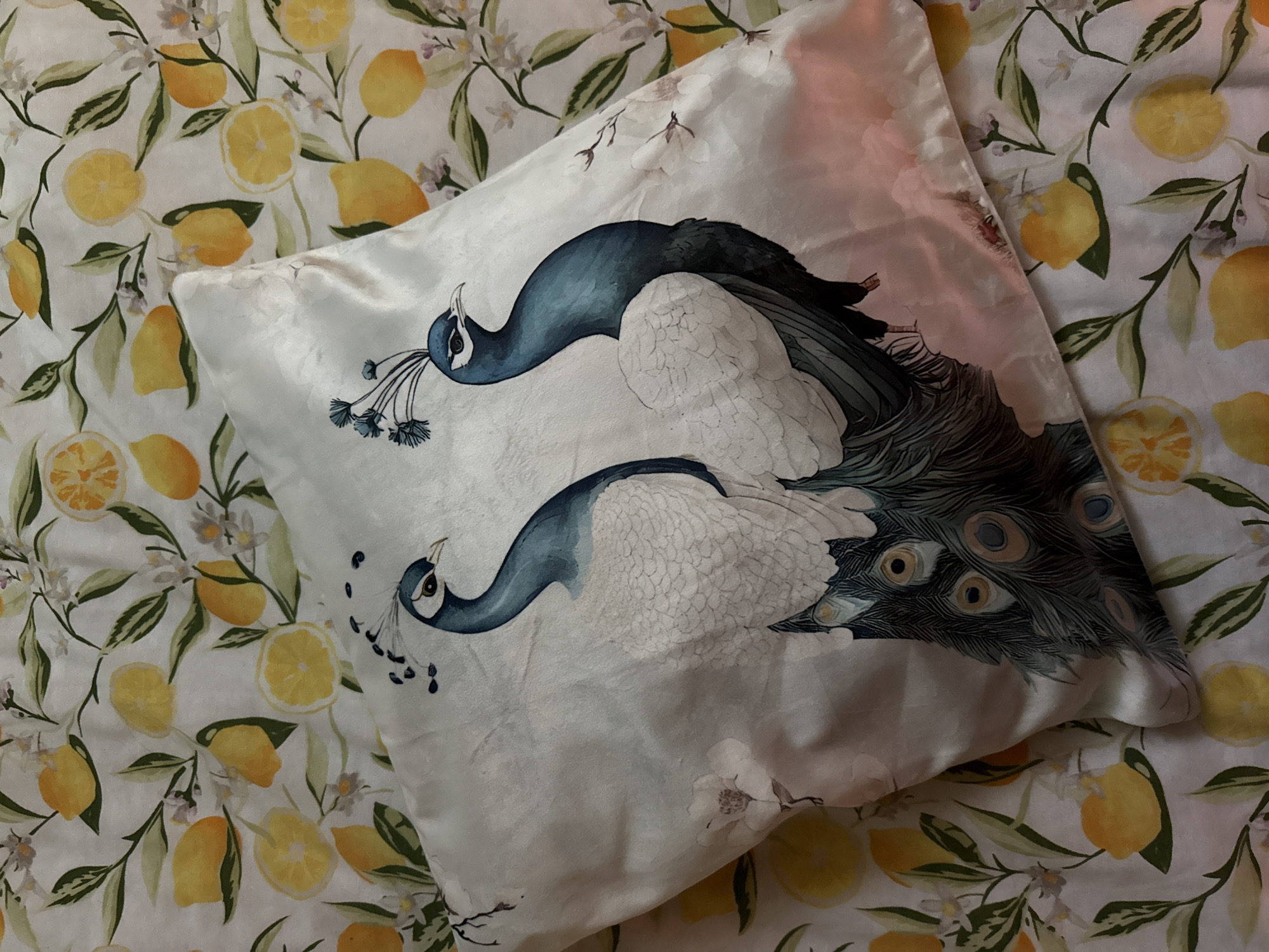 The image shows a white pillow with a peacock design on top of a white bedspread with a lemon and floral pattern. The scene does not appear to be in Pleven, Bulgaria or any discernible city.
