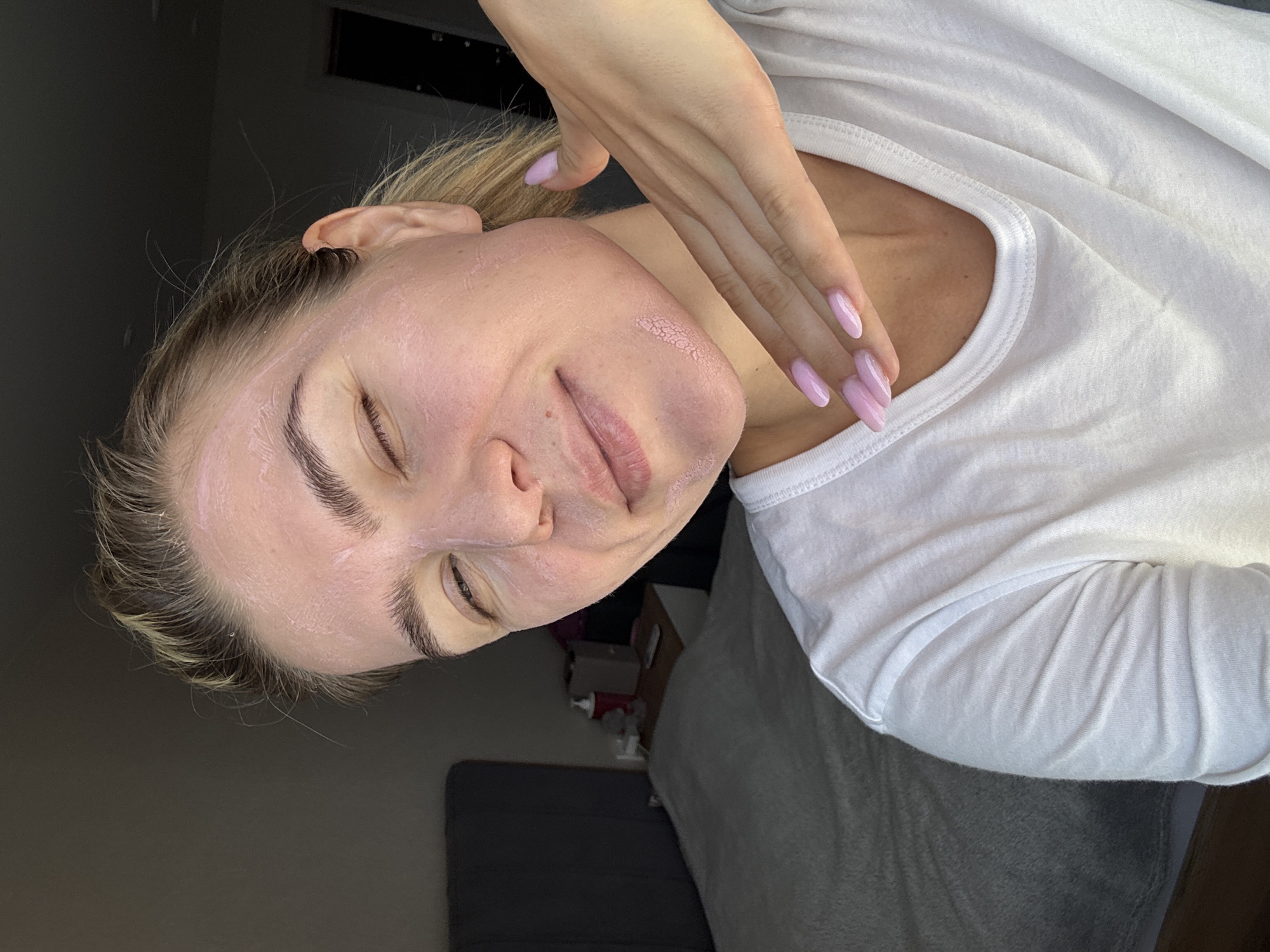 A young woman with blonde hair pulled back is applying a pink facial mask to her face. She is wearing a white t-shirt. Her right hand, with light pink painted nails, is touching her jawline. She is looking at the camera and winking. In the background, a gray headboard and a bed with gray bedding are visible. A nightstand with a red bottle and a gray box is to the right of the bed. The lighting suggests it is daytime, possibly indoors.