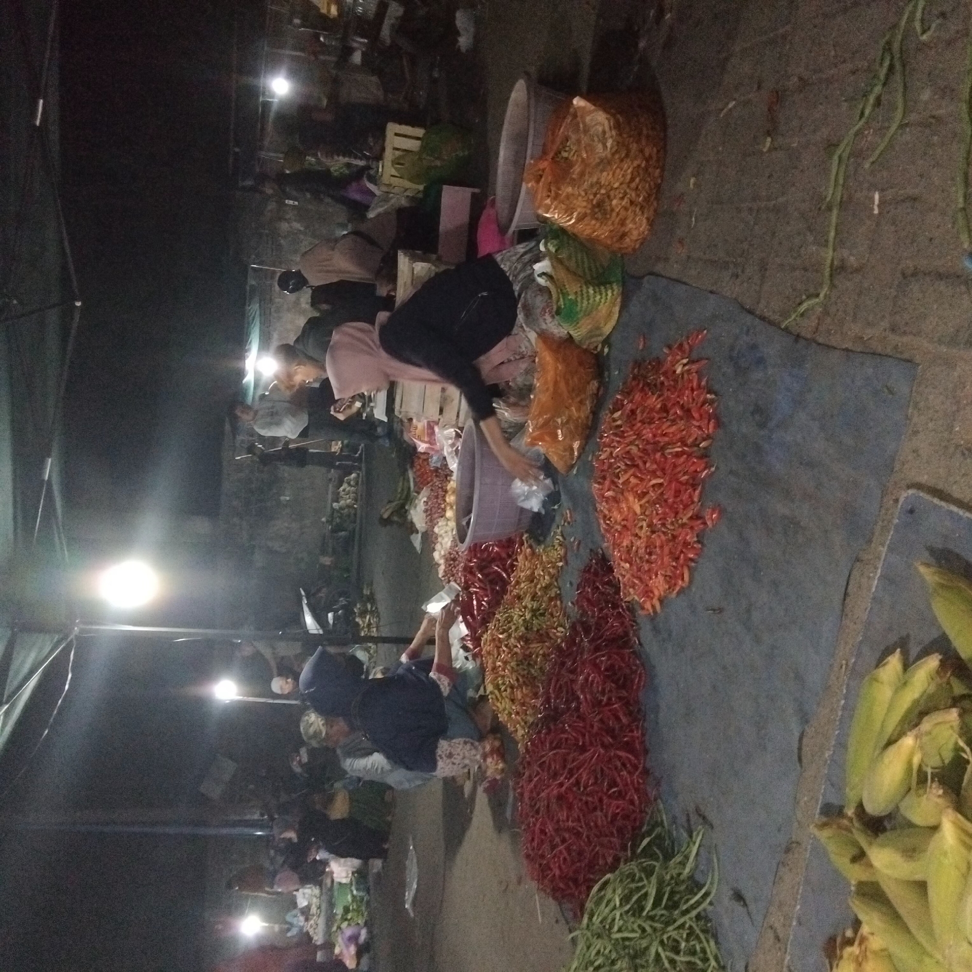 The image shows a night scene at a local market in Banjarbaru, Indonesia. The market appears to be an outdoor or semi-covered space, with tarps providing overhead shelter and bright lights illuminating the scene. Several individuals, likely vendors and customers, are present and engaged in activities related to the sale of produce. In the foreground, there are large piles of various vegetables, including green beans, red chilies, and what appear to be smaller, redder chilies or peppers. Corn is also visible in the lower left corner. People are interacting with the produce, with some sorting or arranging it, while others are possibly making purchases. The overall atmosphere suggests a bustling market environment during nighttime operations.