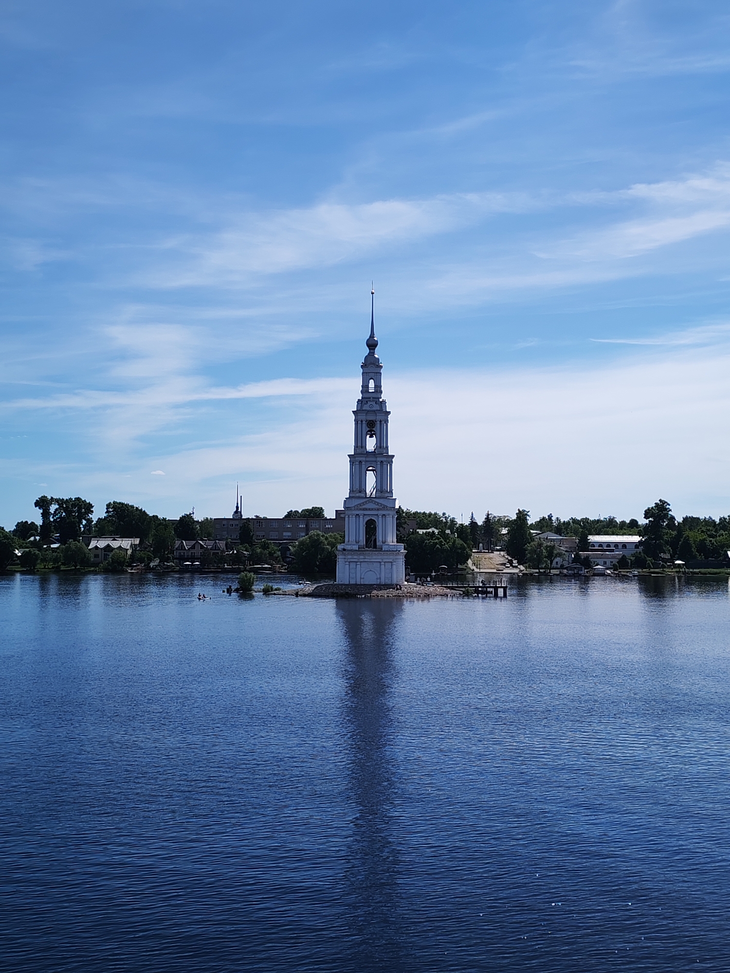 The image displays the iconic Bell Tower of St. Nicholas Cathedral, a prominent white, multi-tiered structure, situated centrally within the Uglich Reservoir in Kalyazin, Russia. The tower occupies a small, partially submerged landmass, with its distinct, elongated reflection visible directly beneath it on the dark blue water.
The sky above is a bright, clear blue, adorned with scattered, wispy white clouds. The water in the foreground and surrounding the tower exhibits small ripples, indicating a calm but not perfectly still surface.
The background reveals the shoreline of Kalyazin, characterized by a mix of dense green trees and various buildings, including residential-style houses and larger, light-colored structures. A long, dark pier or dock extends from the land towards the tower on the right side of the frame. To the left of the tower, two individuals are visible in a small, light-colored boat or kayak on the water. Several more people are discernible on the small landmass around the base of the bell tower and near the dock. A second, smaller boat is partially visible near the dock on the right. The overall scene suggests a clear, sunny day.