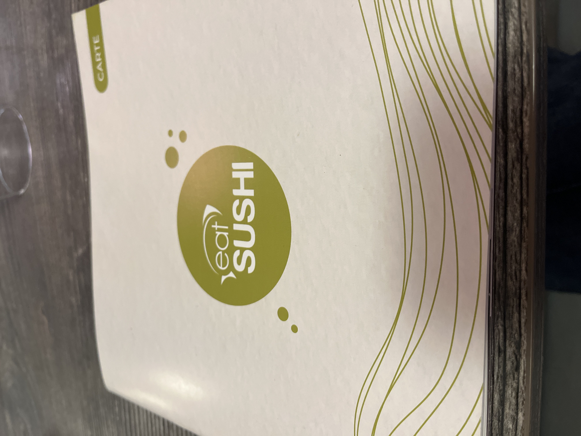 The image shows an "Eat Sushi" restaurant menu placed on a dark-colored table. The menu, oriented vertically from the viewer's perspective and photographed from a slightly overhead angle, features an off-white background. A large, circular lime green logo is centered, containing the white text "Eat SUSHI." The word "Eat" is stylized and positioned above "SUSHI." Small, lime green dots are scattered around the main logo.
In the upper left corner, a vertical lime green tab displays the word "CARTE," which means "menu" in French. The right side of the menu cover is decorated with several thin, wavy lime green lines. Part of a clear drinking glass is visible on the table surface to the left of the menu. The table itself has a dark, textured appearance, possibly wood or laminate. The scene suggests an indoor dining setting, likely a sushi restaurant in Andrézieux-Bouthéon, France, where the menu is presented for ordering. No people or specific activity beyond the presence of the menu are depicted. The time of day and weather are not discernible.