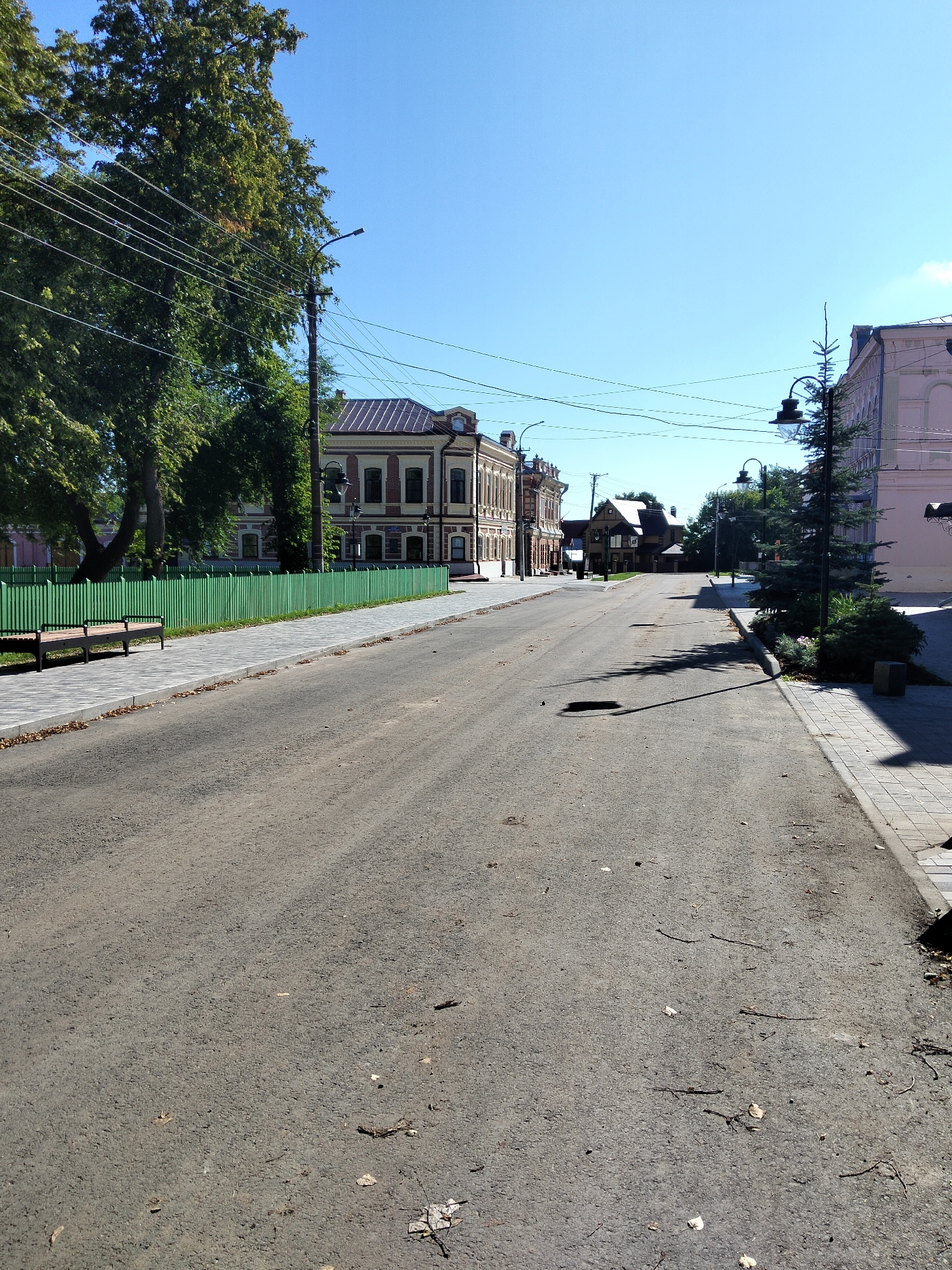 The image depicts a street scene in the city of Kozmodemyansk, Russia. A paved road dominates the foreground, with a sidewalk running along the right side. On the left side of the road, a green wooden fence encloses an area with trees and two benches. Behind the fence, a two-story building with a red-tiled roof and yellow facade is visible. Further down the street, more buildings line both sides, and power lines crisscross overhead. A street lamp with an ornate design stands on the right sidewalk. The sky is clear and blue. The road is somewhat dirty, with scattered leaves and debris.