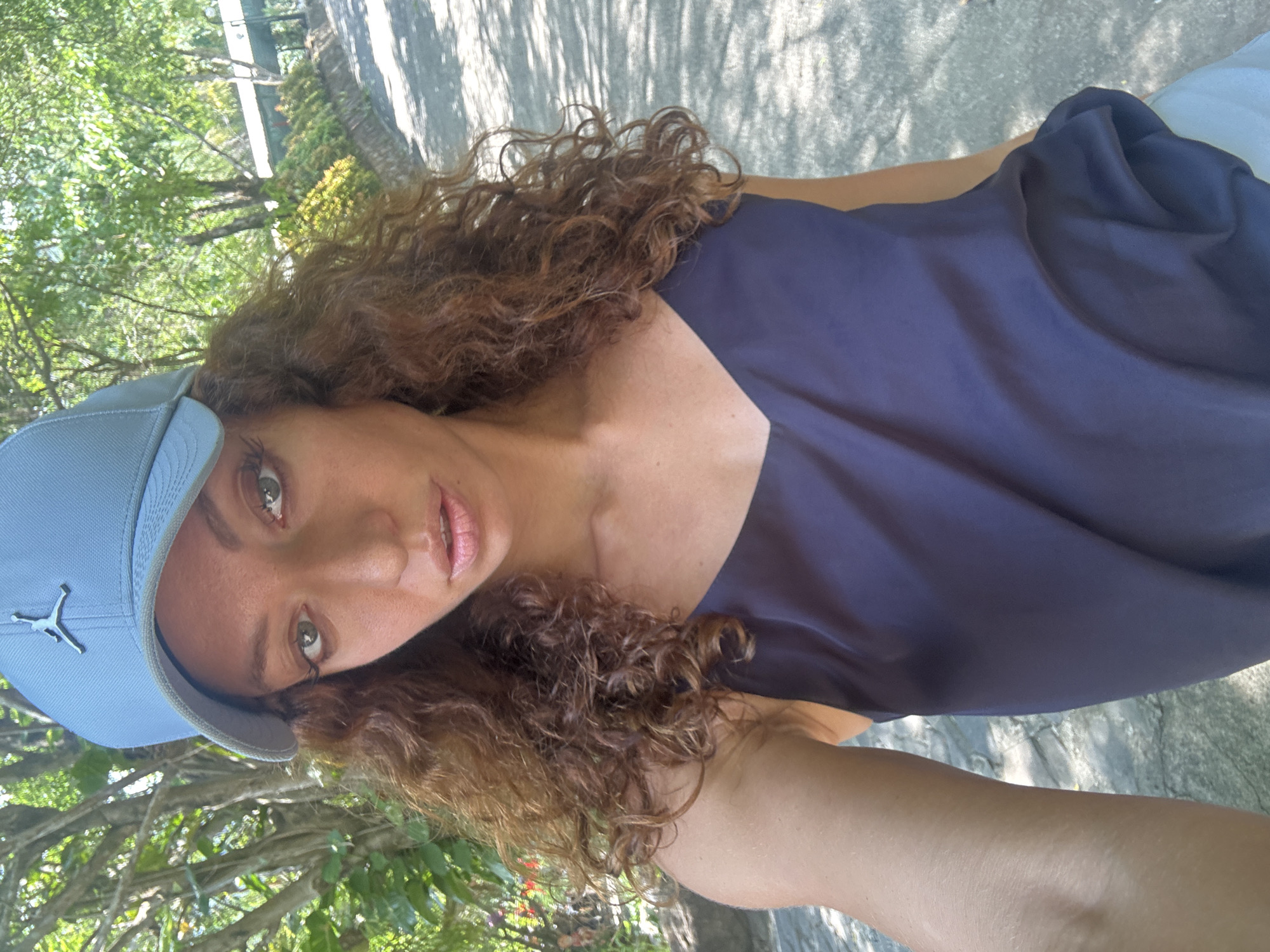 A medium shot depicts a woman with light skin, brown curly hair, and wearing a light blue baseball cap. The cap has the "Jumpman" logo on the front. She is wearing a dark blue top. The woman is outside, with trees and greenery visible in the background. The environment suggests an outdoor park or garden setting. The presence of foliage and dappled sunlight indicates a location with trees. There is no indication from the image that the location is in Đà Nẵng, Vietnam.