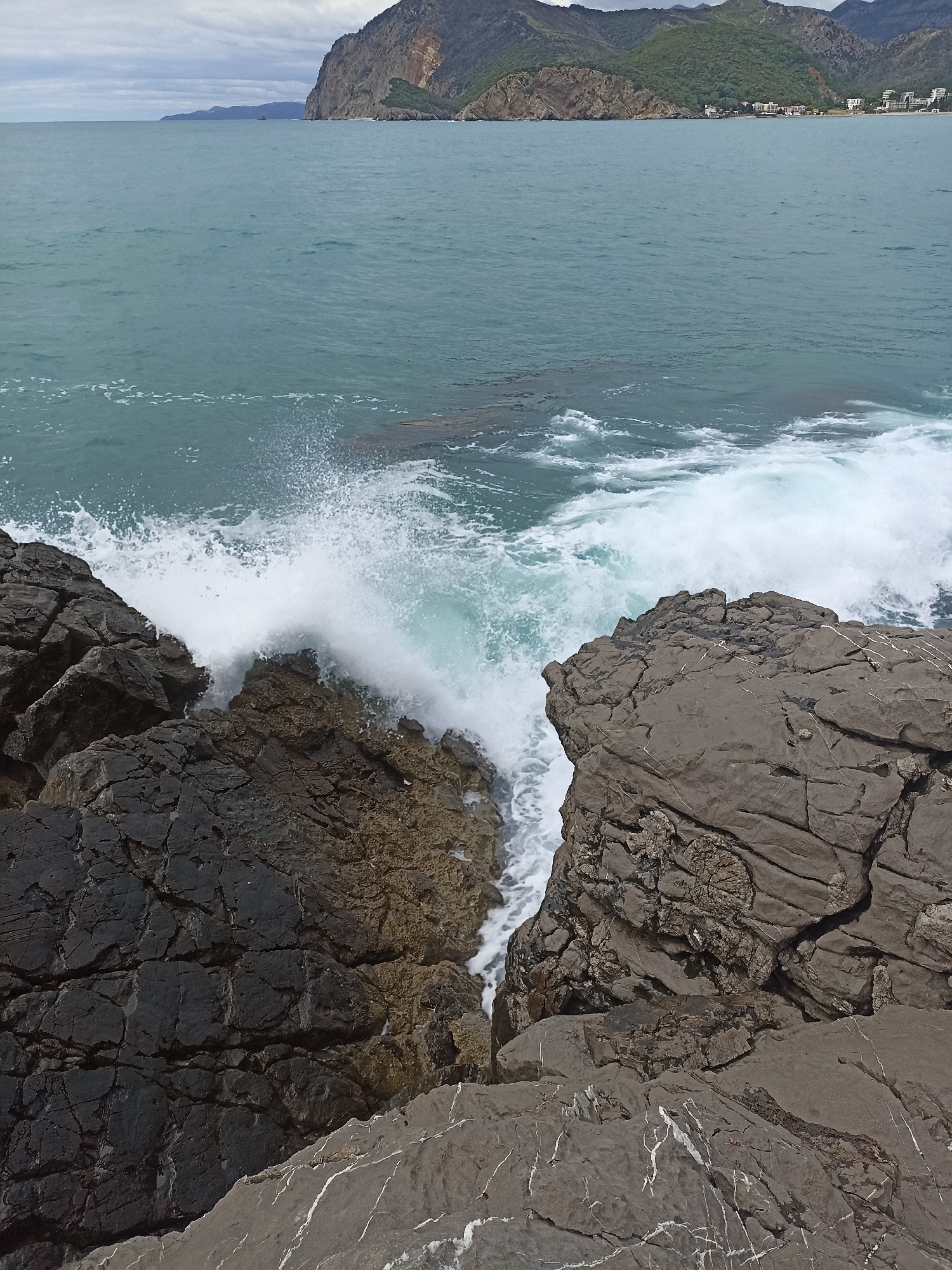 The image shows a view of the sea from a rocky outcropping in Čanj, Montenegro. In the foreground, jagged, dark grey and brown rocks form a small crevice where waves are crashing, creating white foam. In the mid-ground, the sea is a turquoise color with gentle waves further out. In the background, a large, mountainous landmass is visible, covered in green vegetation and some buildings near the shore. The sky is overcast with grey clouds.