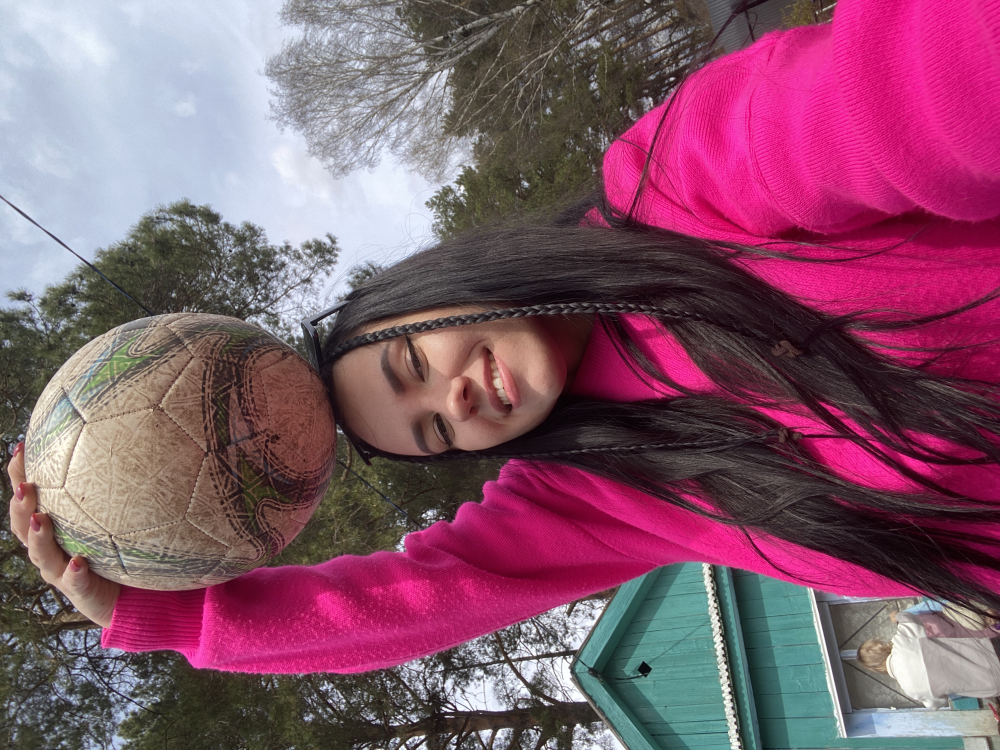 The image features a young woman with long dark hair, styled with small braids, wearing a bright pink knitted sweater. She is smiling broadly and looking towards the camera, holding a distressed-looking soccer ball with a camouflage-like pattern in her right hand, resting it against her head. Her nails are painted a light red or pink.
The setting appears to be outdoors, with a cloudy sky visible overhead, indicating an overcast day. In the background, there are trees, some appearing bare, suggesting late winter or early spring, and others are evergreens. Partially visible on the right side of the frame is a building with teal or light green wooden siding and white decorative trim. Through a window of this building, another person, possibly a child, wearing a white garment, is vaguely visible. The image is taken from a low-angle perspective, looking up. There is no visible text in the image. The scene suggests a casual, playful moment in an outdoor, possibly rural or semi-rural, environment in Chaikovsky, Russia.